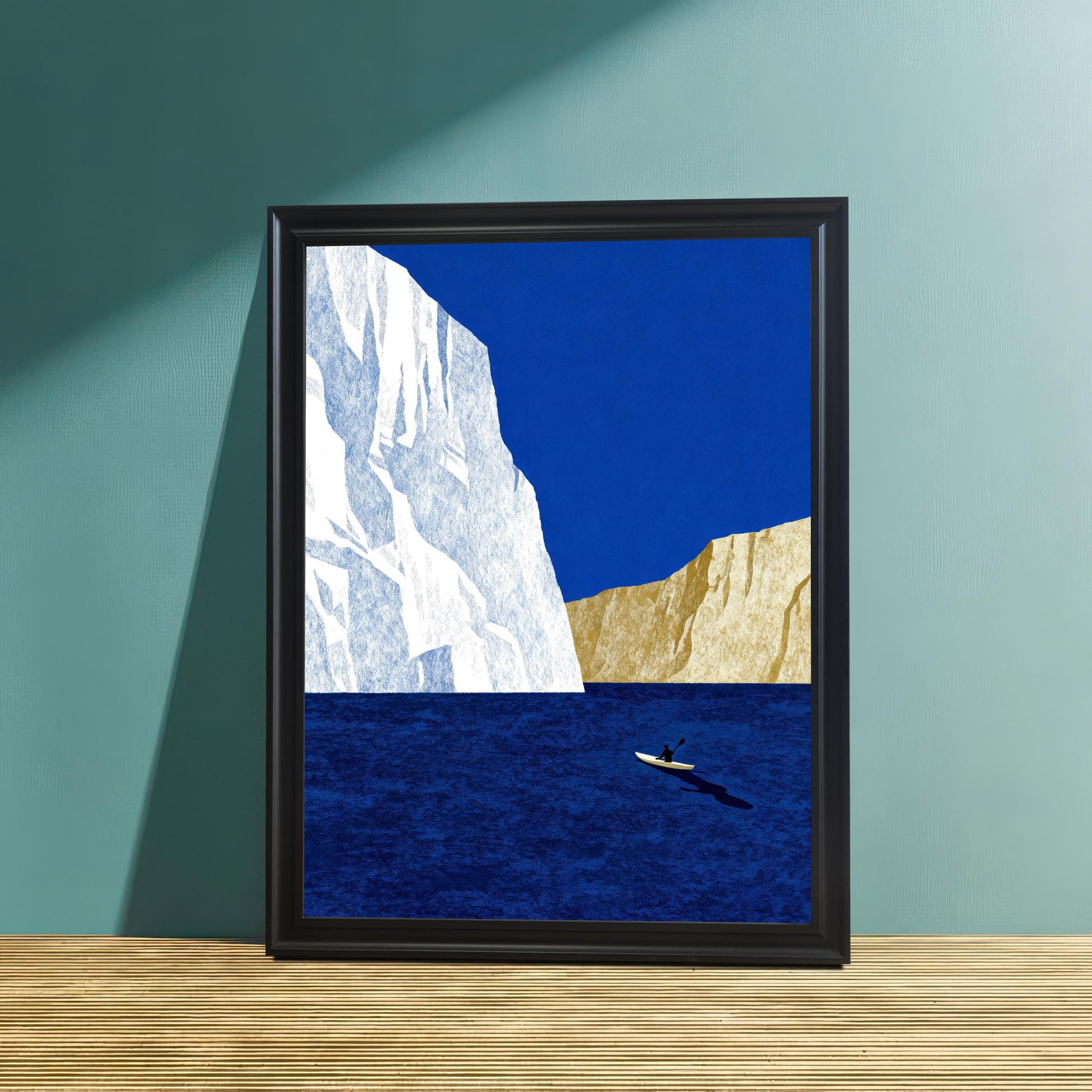 Minimalist iceberg and kayaker art print in a sleek black wood frame, leaning against a textured teal wall on a light wood surface.