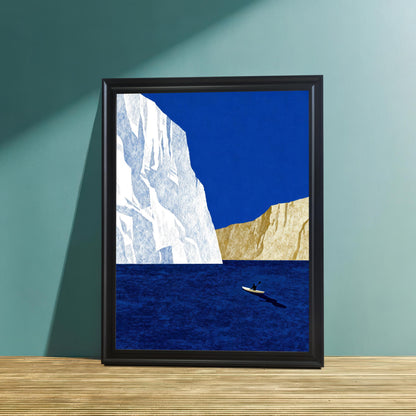 Minimalist iceberg and kayaker art print in a sleek black wood frame, leaning against a textured teal wall on a light wood surface.