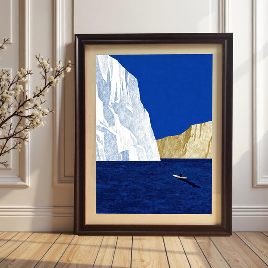 Large-scale seascape art print in a coffee-colored wood frame with a cream mat board, leaning on a light wood floor against a white paneled wall.
