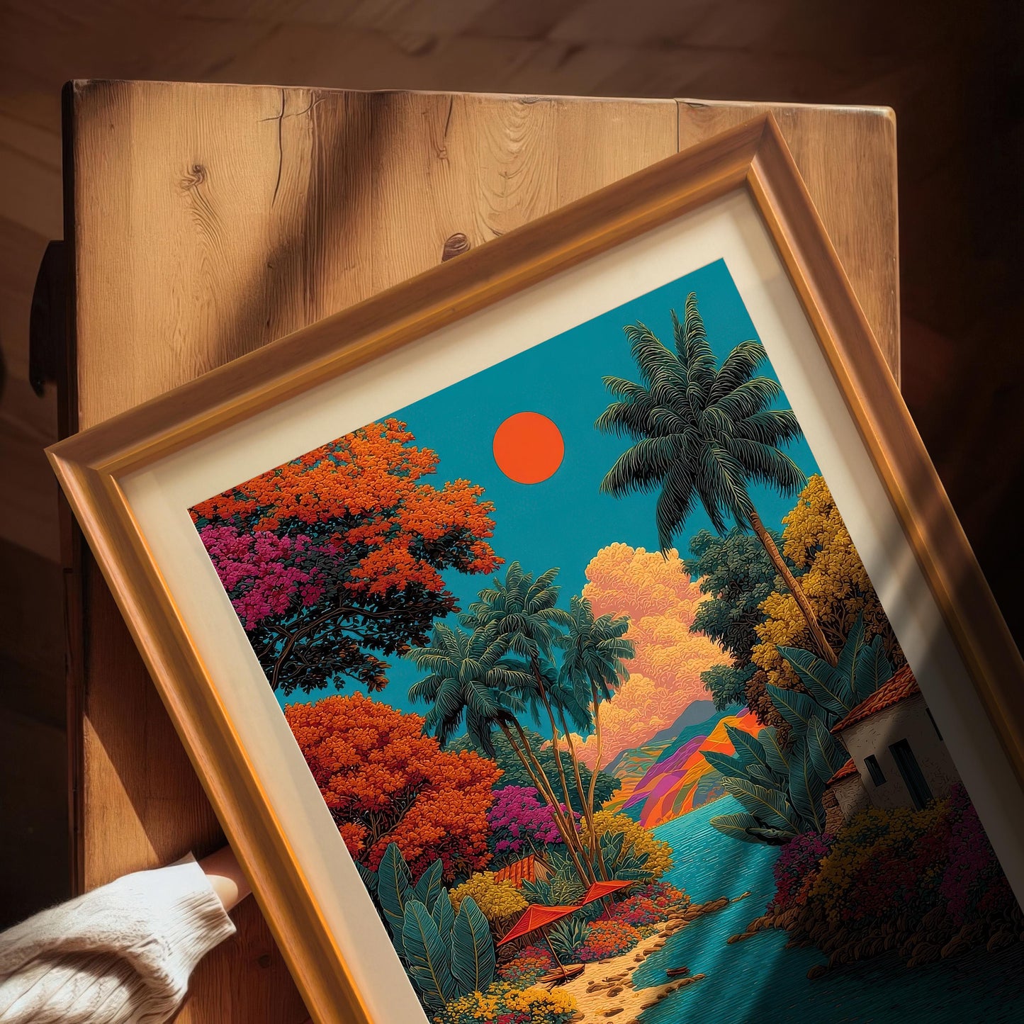 Overhead close-up view of the vibrant tropical landscape art print in an Orange frame with a mount, resting on a wooden surface. A hand in a cream sweater is visible in the corner.