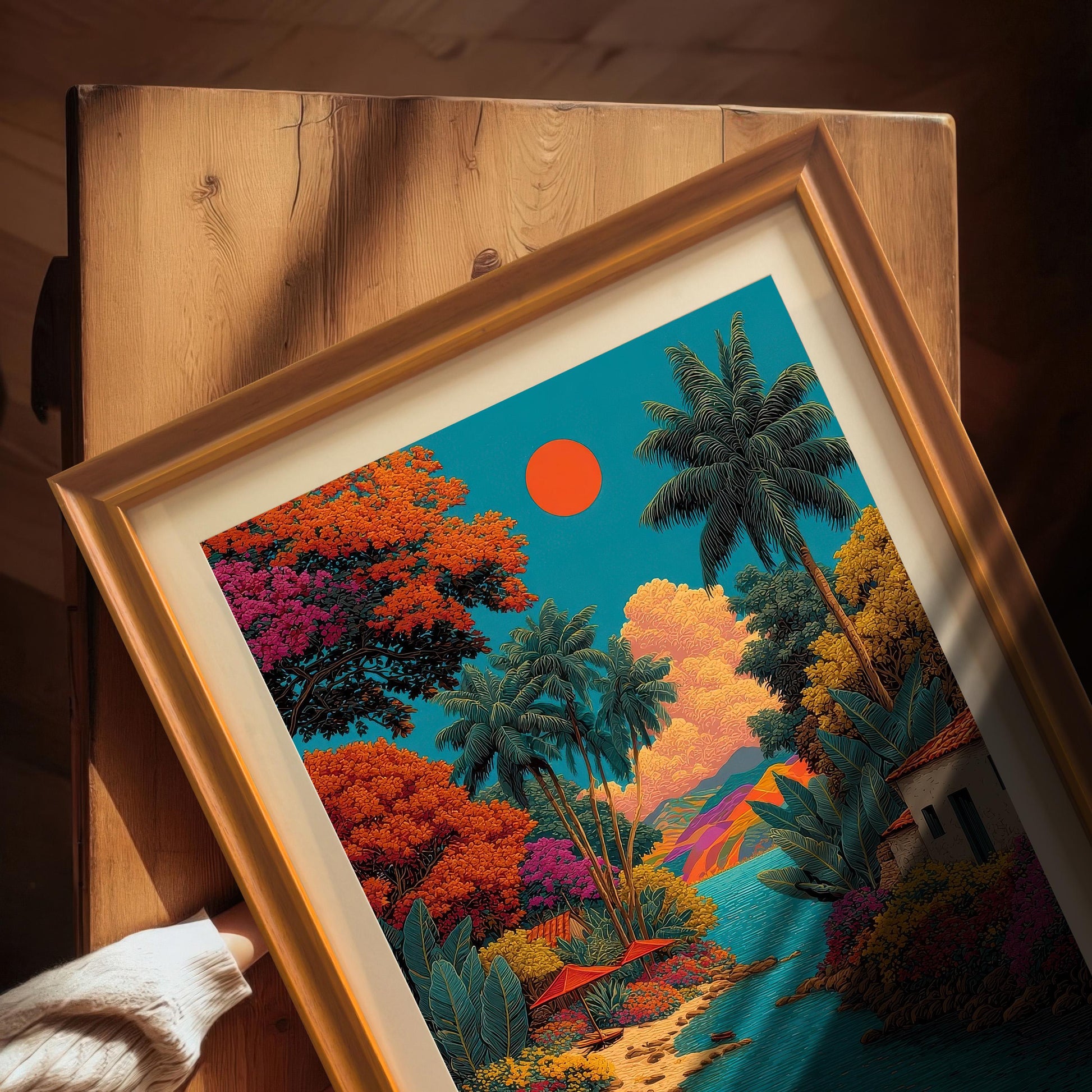 Overhead close-up view of the vibrant tropical landscape art print in an Orange frame with a mount, resting on a wooden surface. A hand in a cream sweater is visible in the corner.