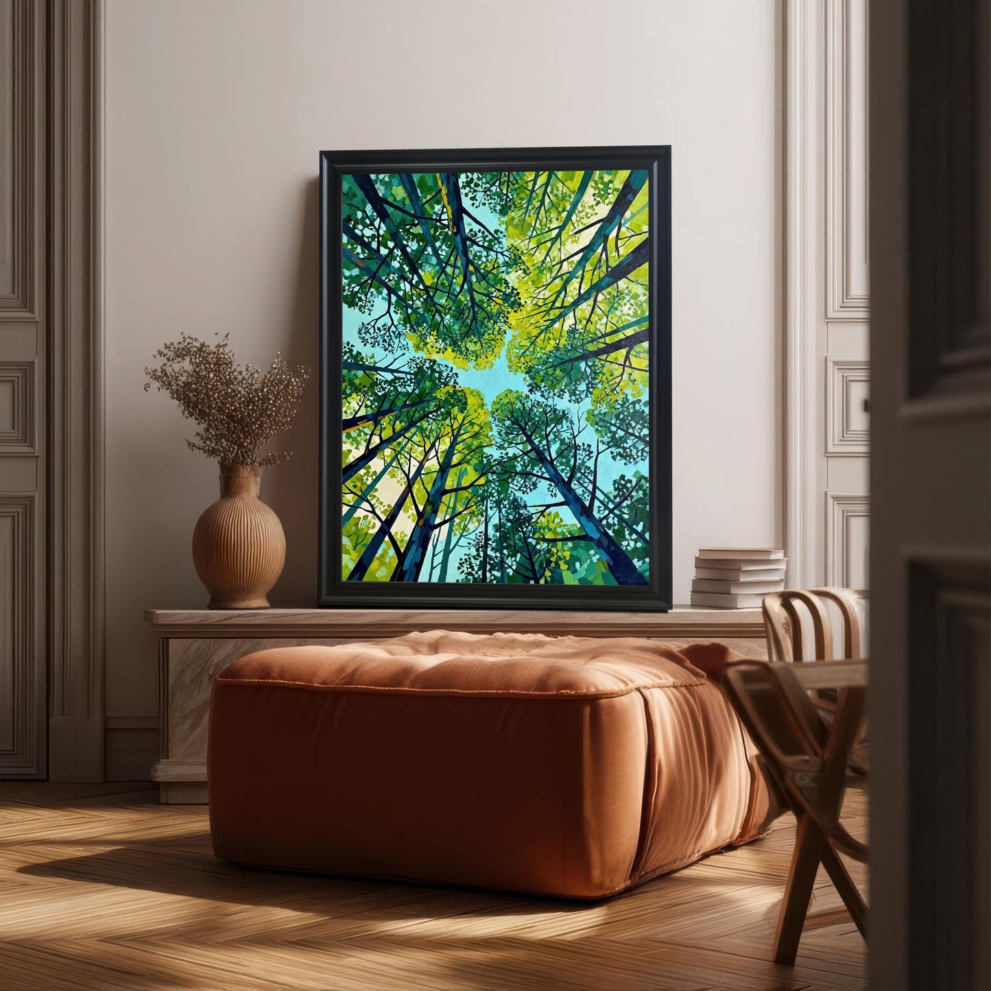 Large black-framed forest canopy art print styled in a bright modern room, leaning on a marble console table next to a large orange ottoman and a terracotta vase.