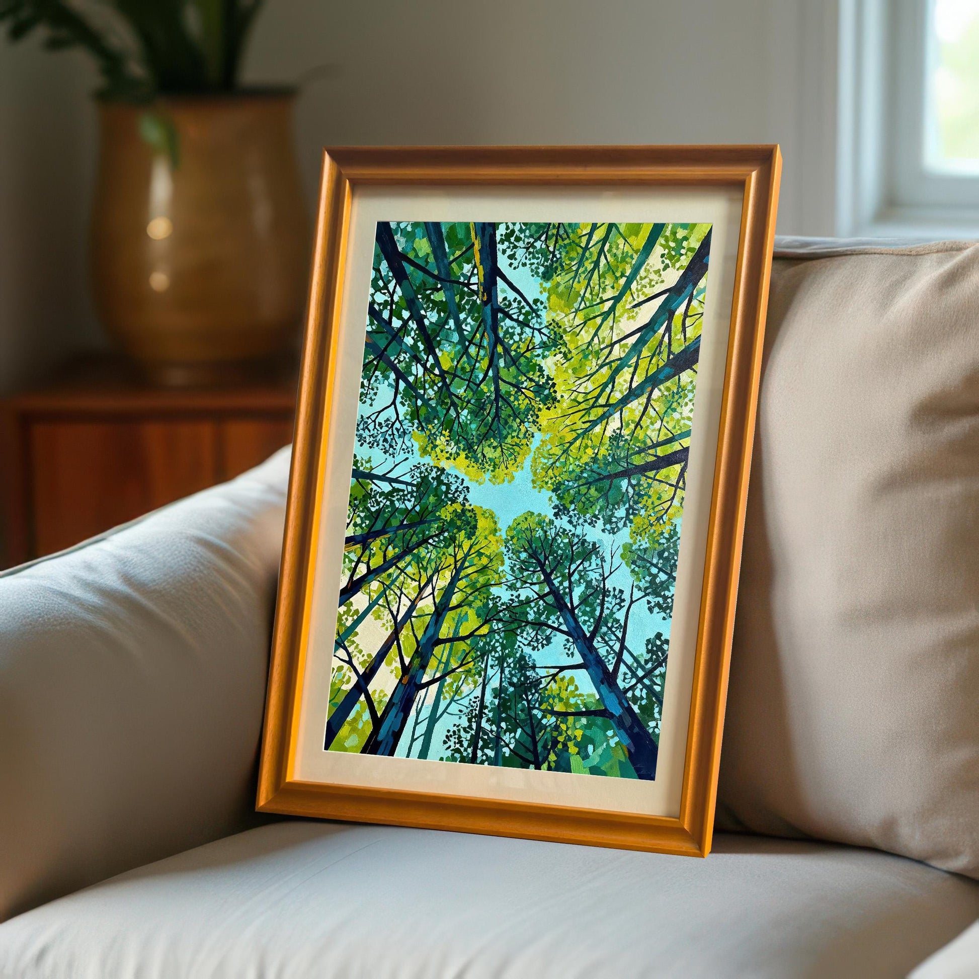 A matted green forest art print in an orange wood frame resting on a grey fabric sofa in a bright, modern interior.