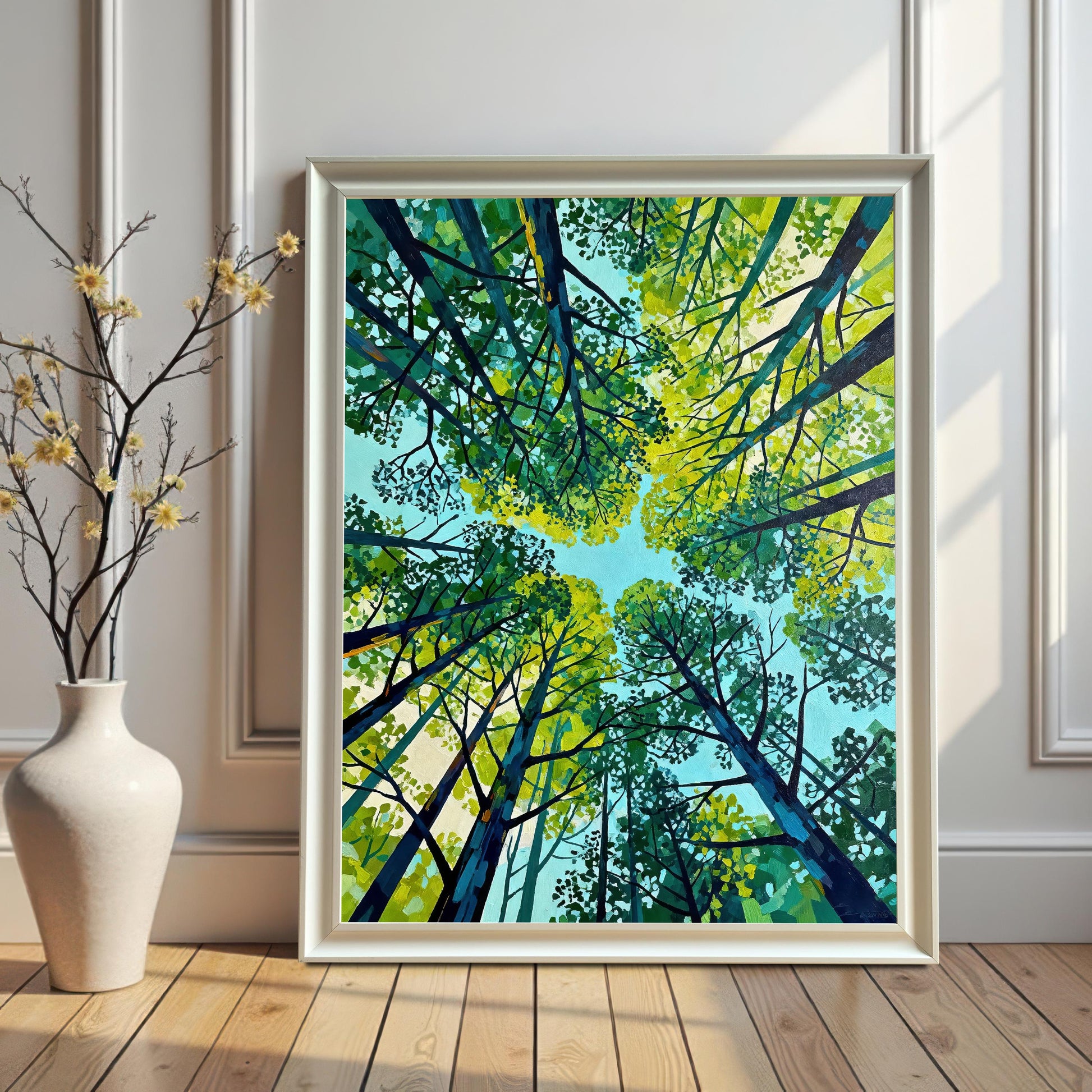 Vertical green tree art print in a sleek white wood frame, leaning against a paneled white wall next to a white ceramic vase with delicate yellow flowers.