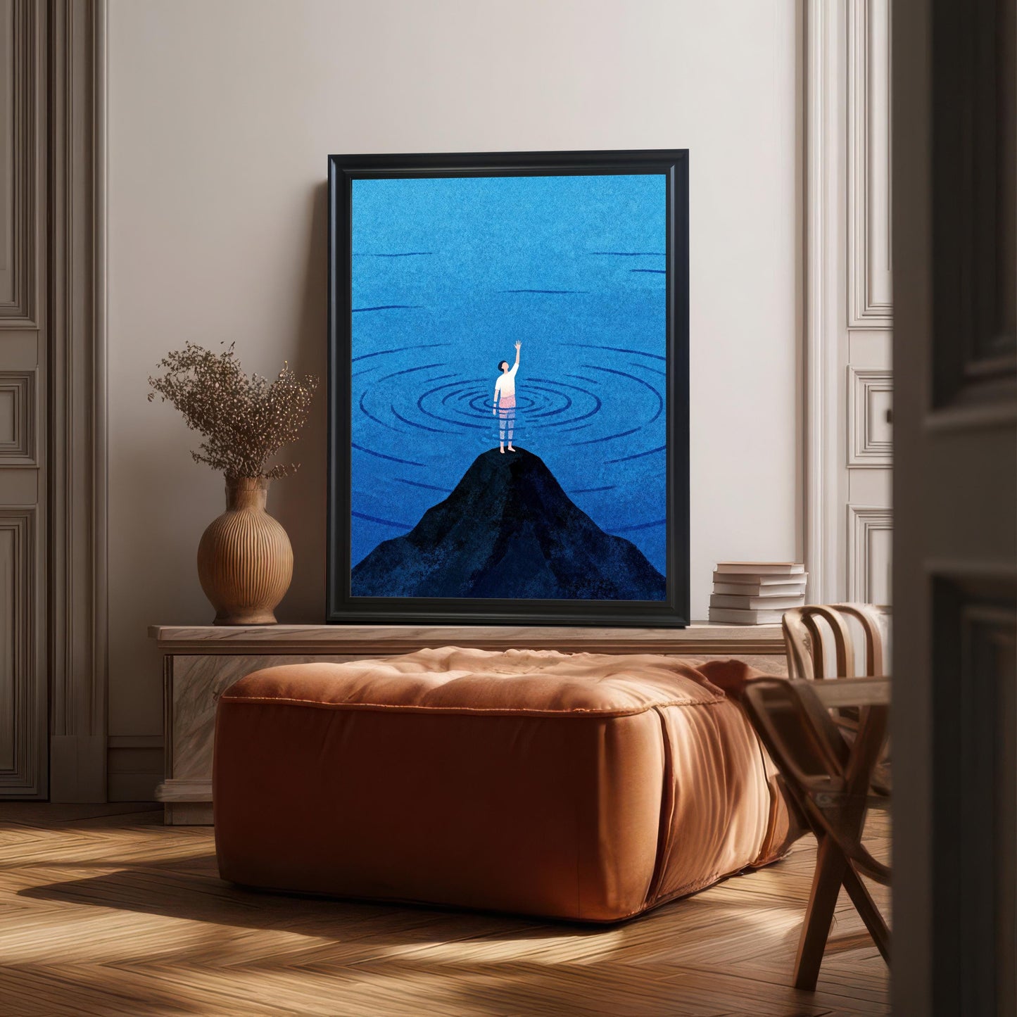 A sleek black-framed art print without a mount, displayed in a sophisticated living room setting. The frame sits on a low wooden console next to a textured clay vase with dried flowers and a stack of books, with a large cognac-colored velvet ottoman in the foreground and sunlight streaming across the parquet floor.