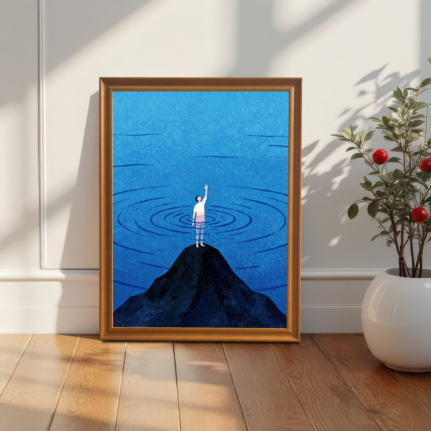 An orange-toned wood frame containing a minimalist blue sea print, standing on a light wood floor next to a white ceramic vase with a green plant. Soft sunlight and shadows fall across the white wall behind it.