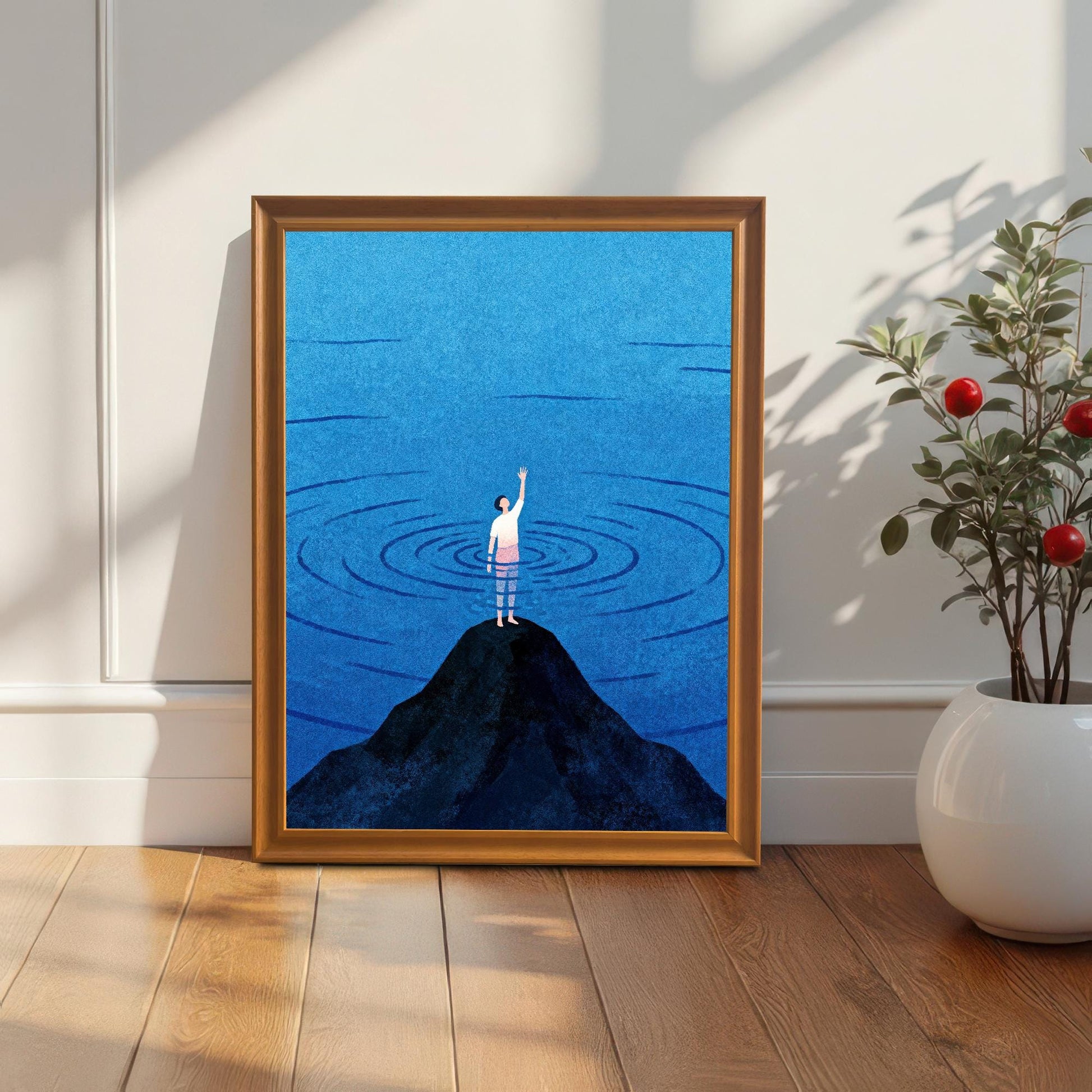 An orange-toned wood frame containing a minimalist blue sea print, standing on a light wood floor next to a white ceramic vase with a green plant. Soft sunlight and shadows fall across the white wall behind it.