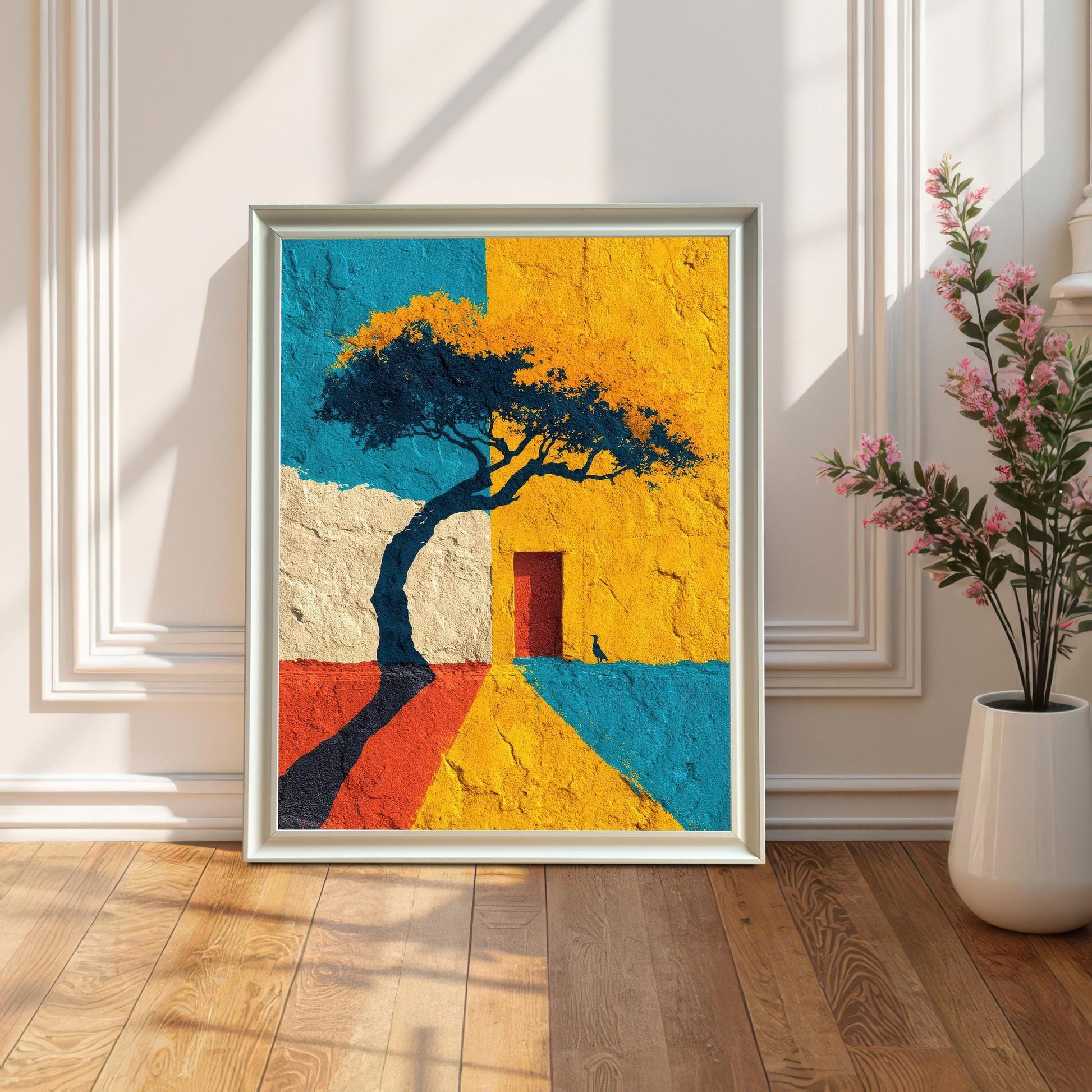 A framed art print in a white frame, leaning on a wooden floor against a white paneled wall (wainscoting). The art is brightly colored and features a tree silhouette, a red door, and blocks of yellow, blue, and red texture. Sunlight casts long, dramatic shadows across the floor. A vase with pink flowers sits to the right.