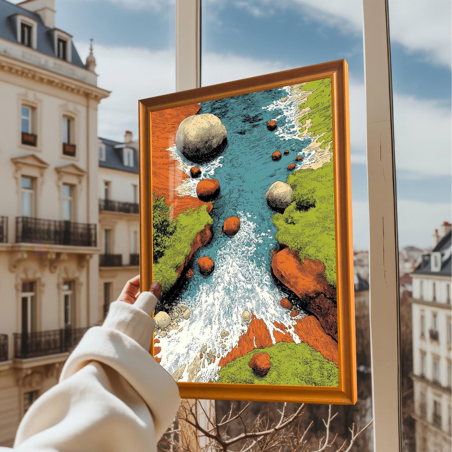A hand holding up the framed art print (in an Orange/light wood frame) in front of an apartment window overlooking a cityscape on a sunny day. The art is a vibrant, textured illustration of a rocky river.