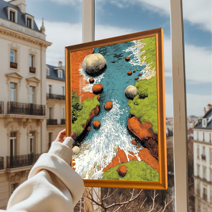 A hand holding up the framed art print (in an Orange/light wood frame) in front of an apartment window overlooking a cityscape on a sunny day. The art is a vibrant, textured illustration of a rocky river.
