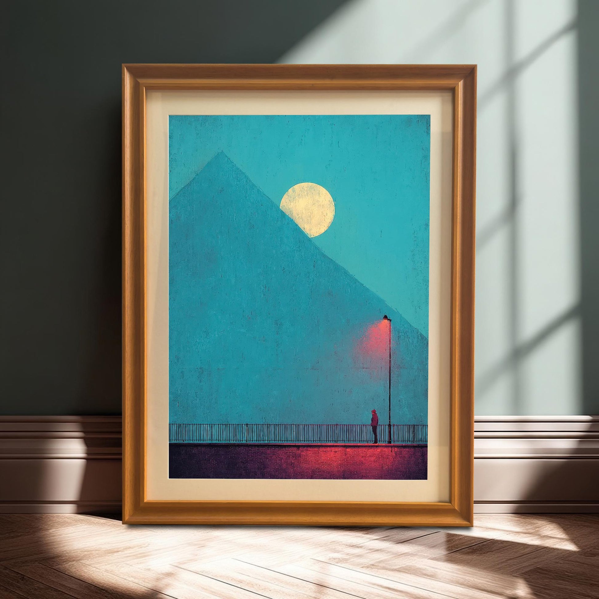 A framed art print with an orange/light wood frame and a mount/matting, leaning against a dark teal wall on a wooden floor with dramatic shadows. The art features the moody pyramid/moon scene.