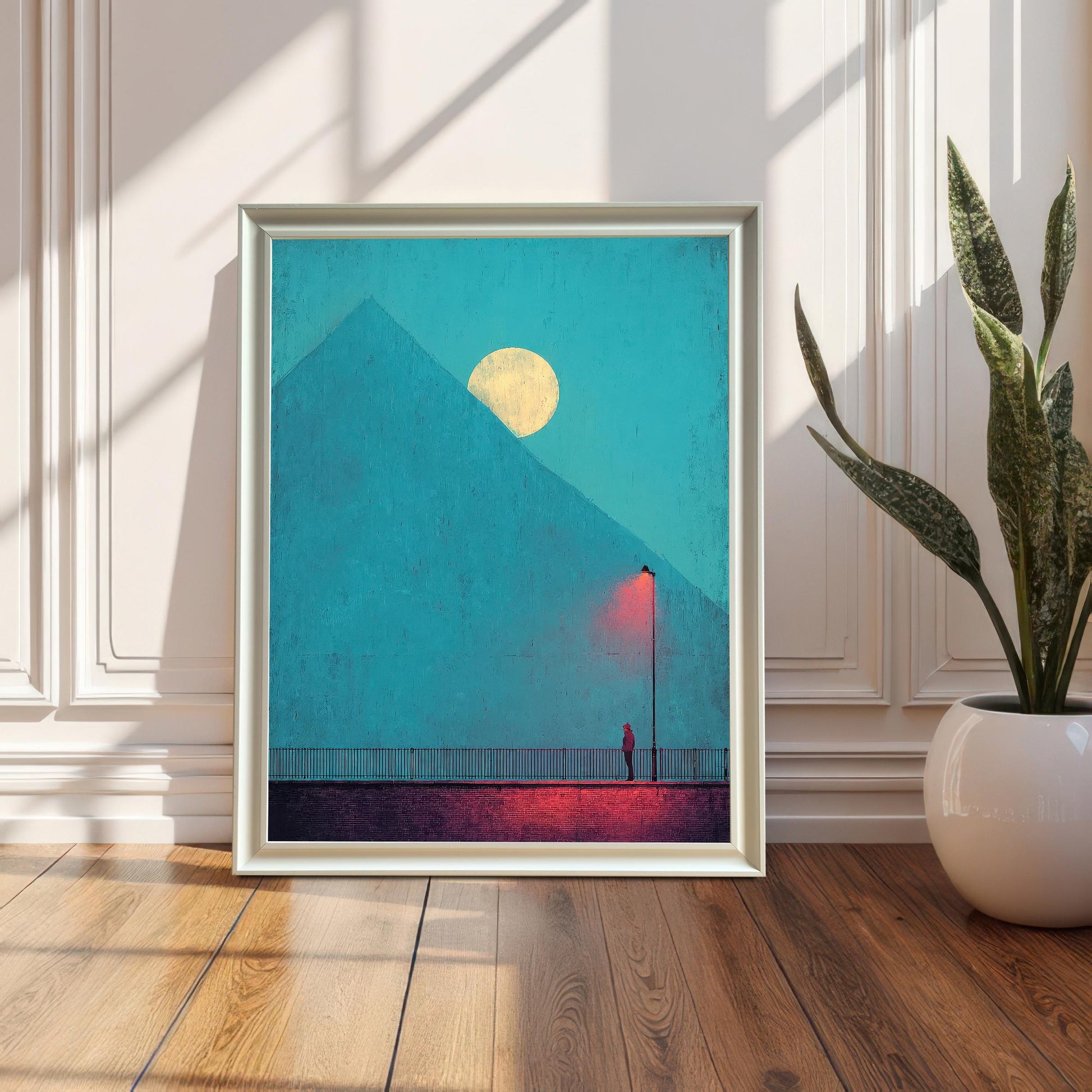 A framed art print in a white frame, leaning on a wooden floor against a white paneled wall (wainscoting). The artwork is a moody, minimalistic scene with a figure under a red lamp post and a yellow moon behind a blue pyramid/mountain. Sunlight casts long, dramatic shadows. A potted snake plant sits to the right.