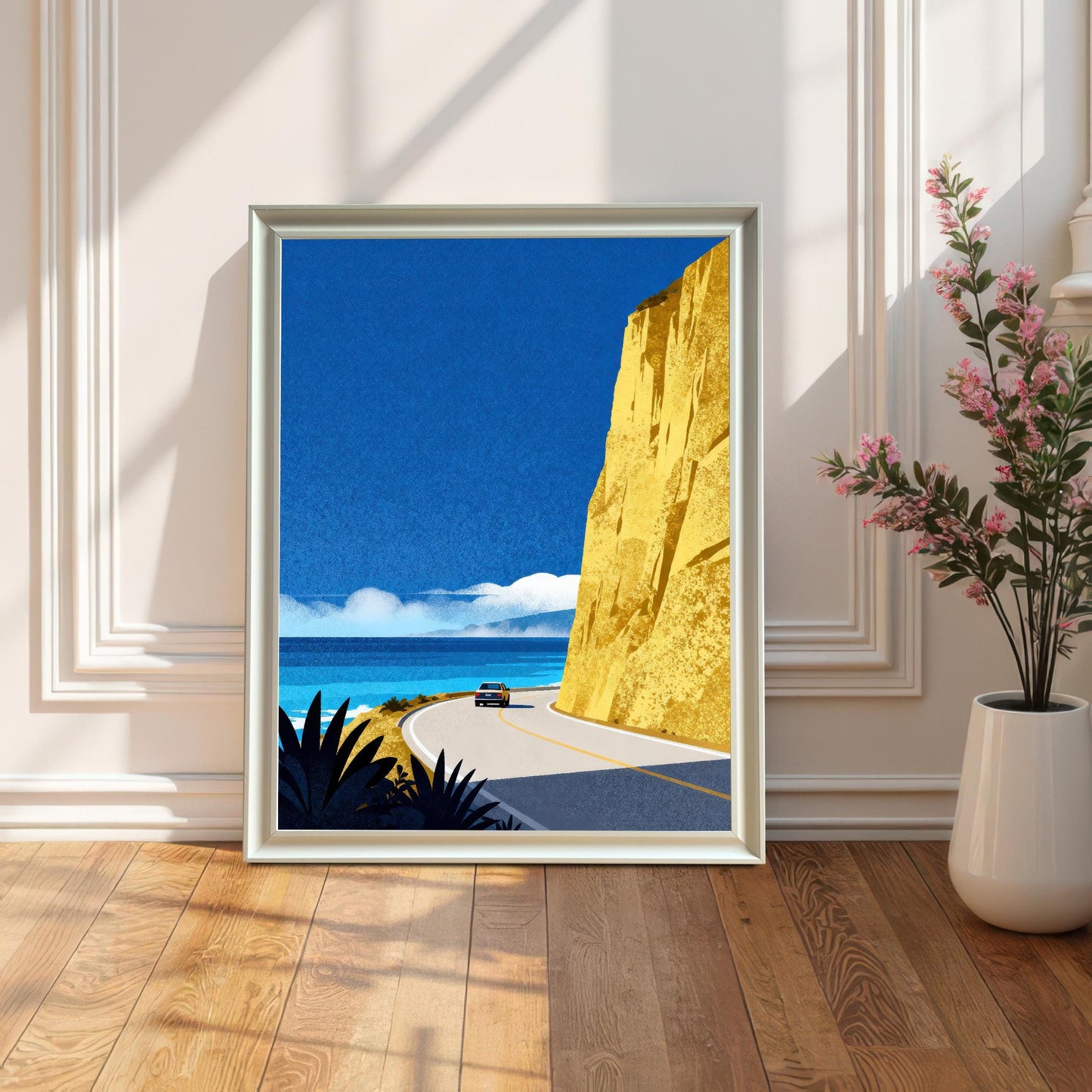 A white wood-framed art print leaning on a polished wooden floor against a white paneled wall, placed next to a white vase with pink flowering branches.