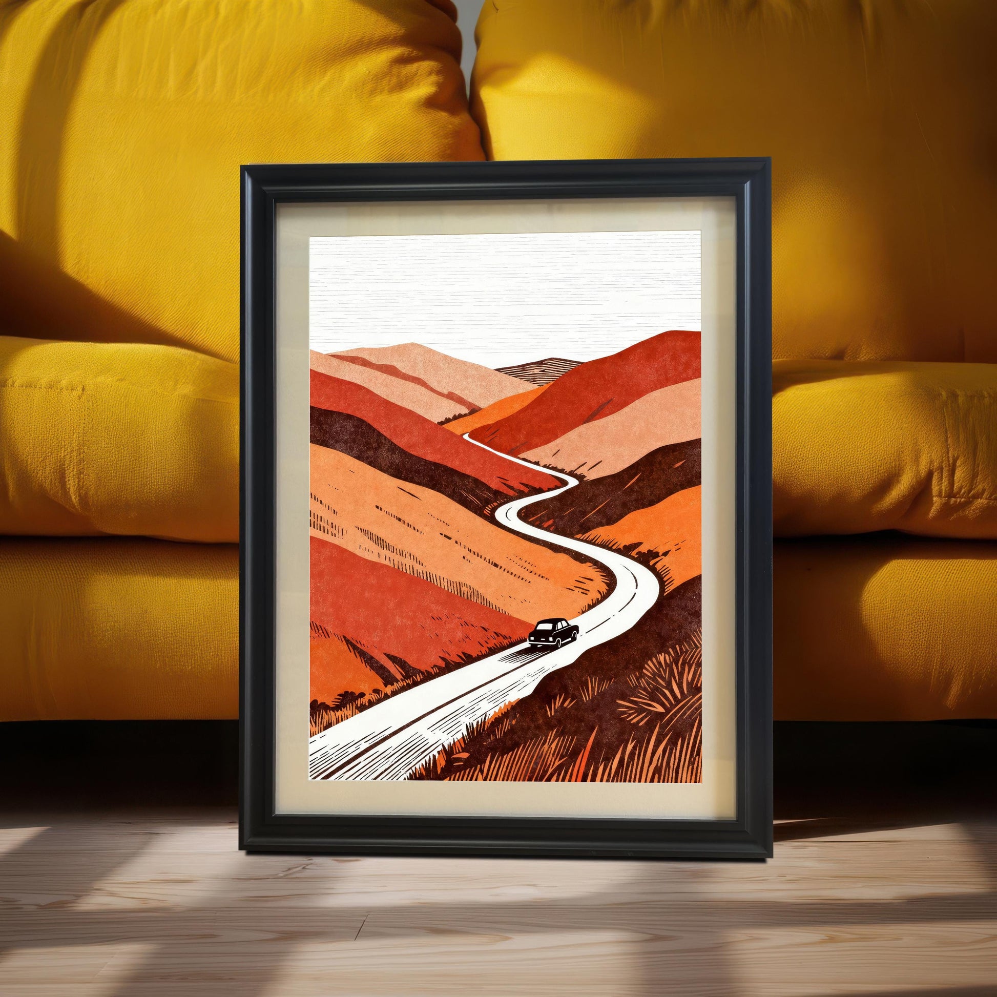 A black wood-framed landscape art print of a winding road through orange hills, leaning against a bright yellow sofa in a sunlit room.