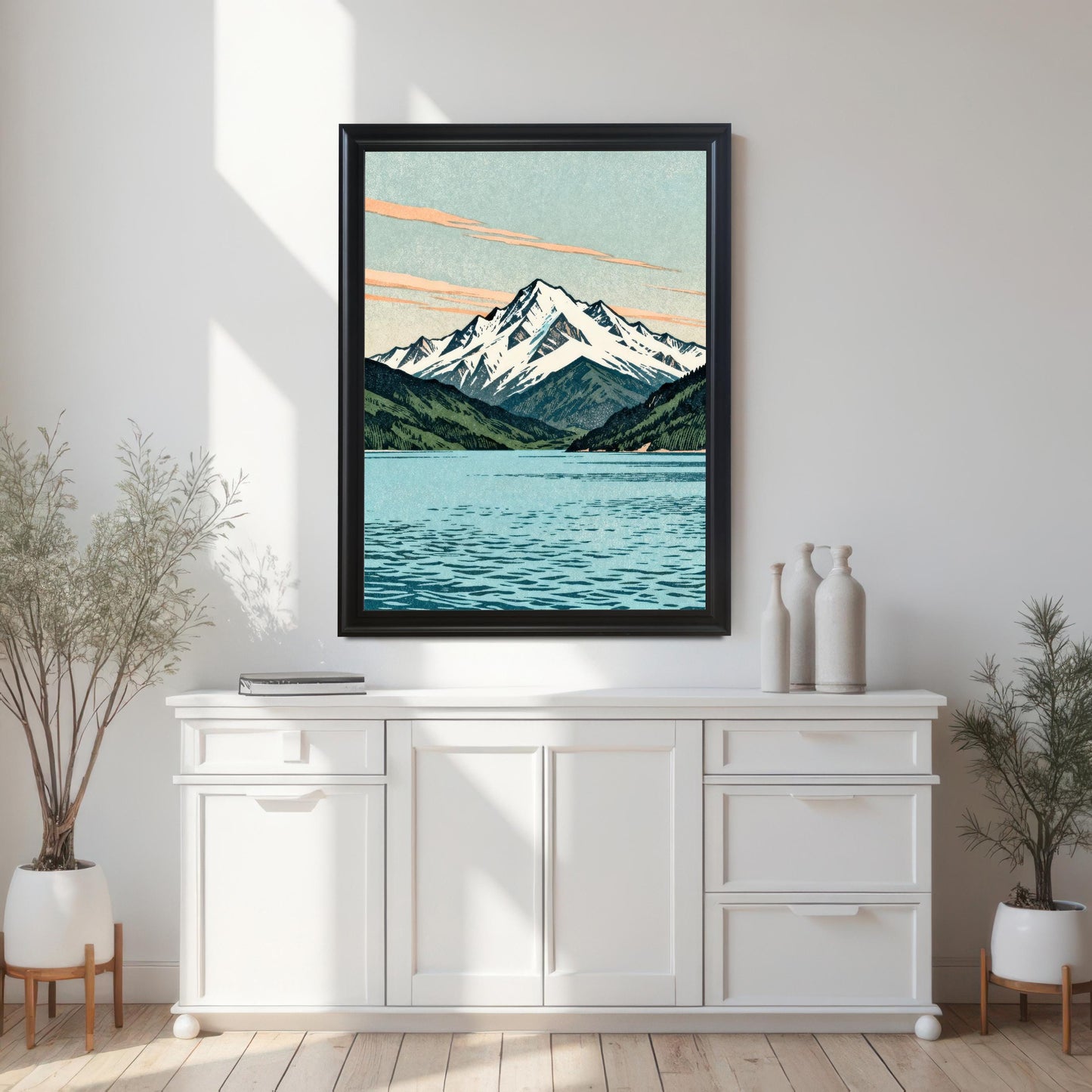 A large black wood framed mountain art print without a mount, hanging on a minimalist white wall above a modern white sideboard cabinet with decorative ceramic vases and potted plants.