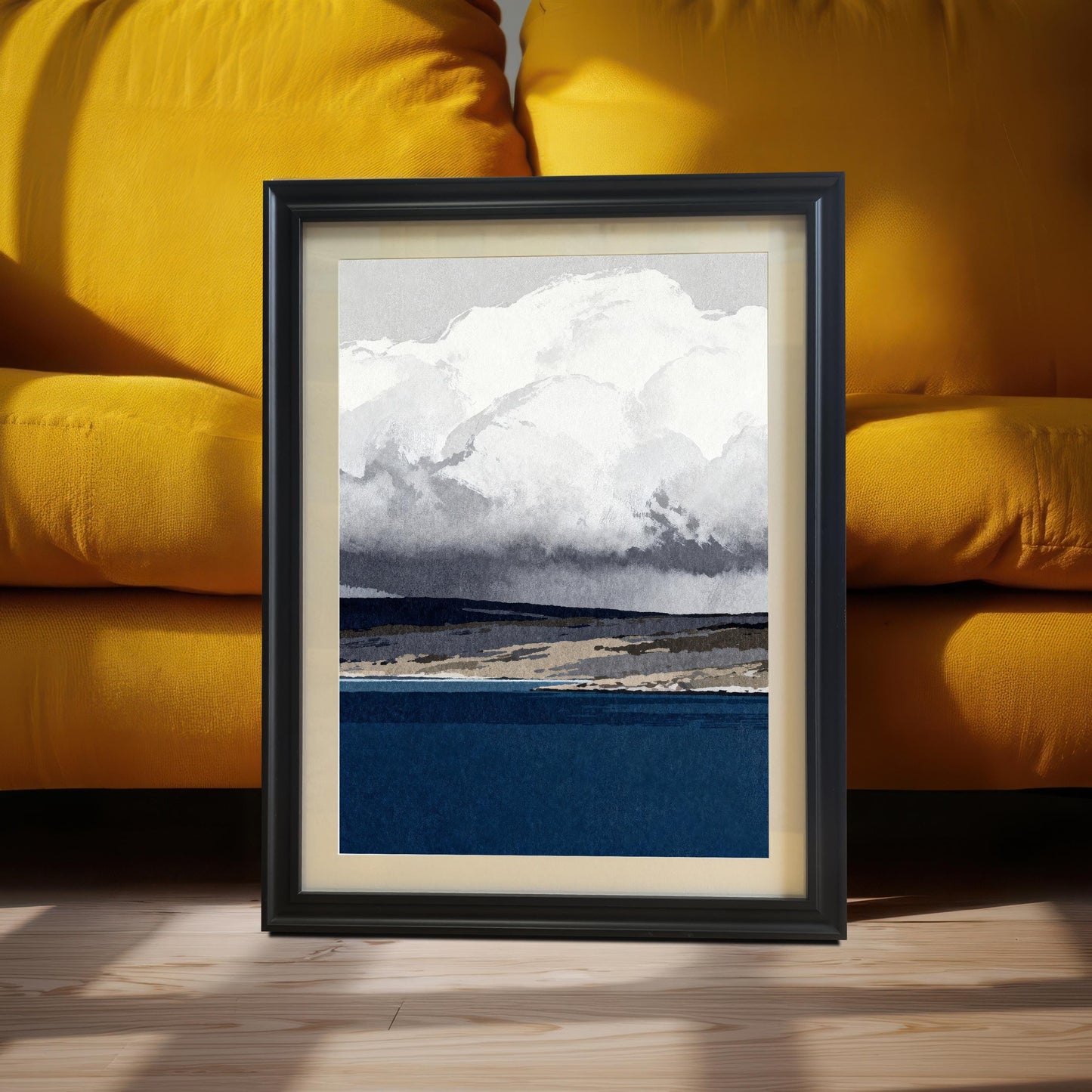 A black wood-framed abstract landscape print featuring a deep blue sea and large white clouds, leaning against a vibrant yellow sofa in a brightly lit room.