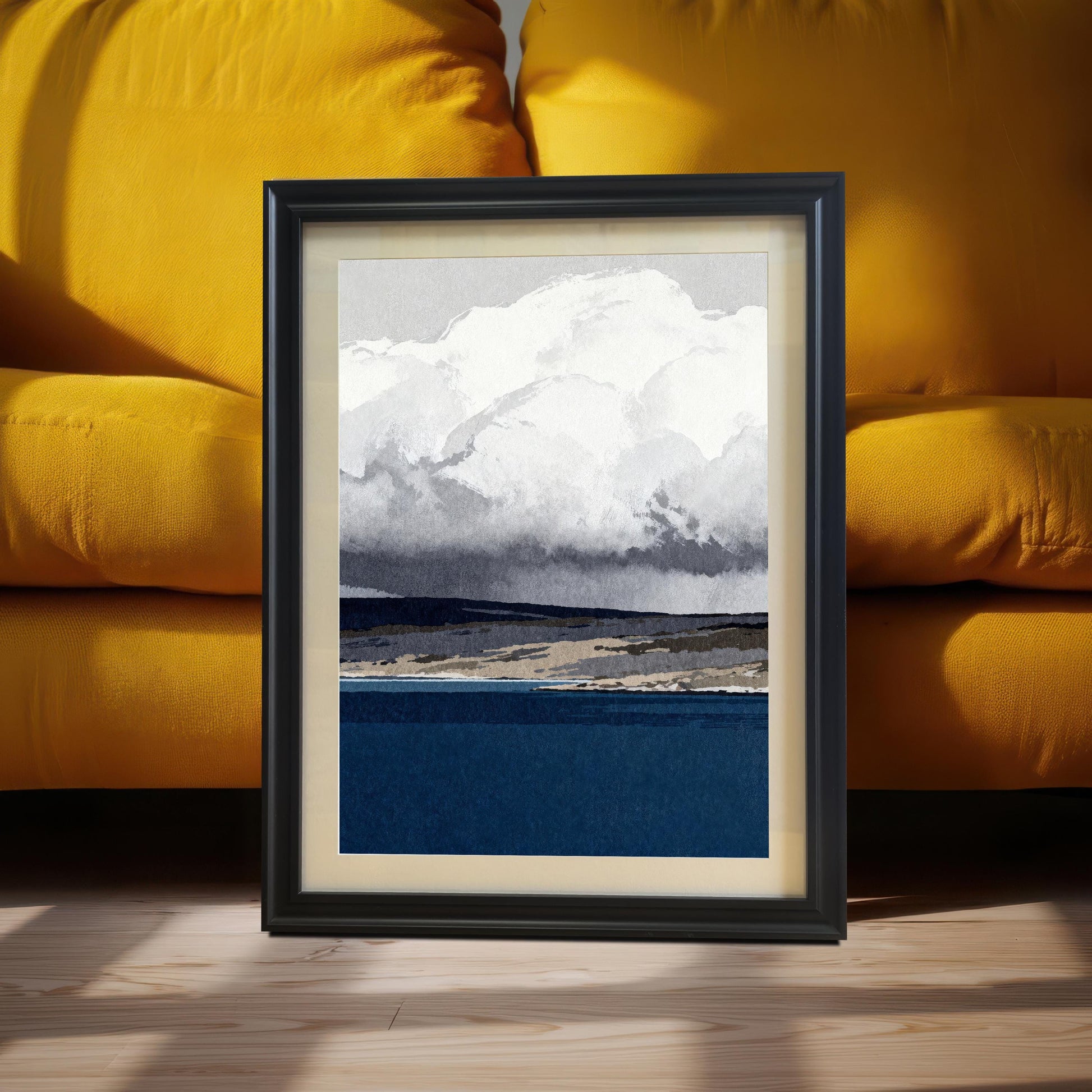 A black wood-framed abstract landscape print featuring a deep blue sea and large white clouds, leaning against a vibrant yellow sofa in a brightly lit room.