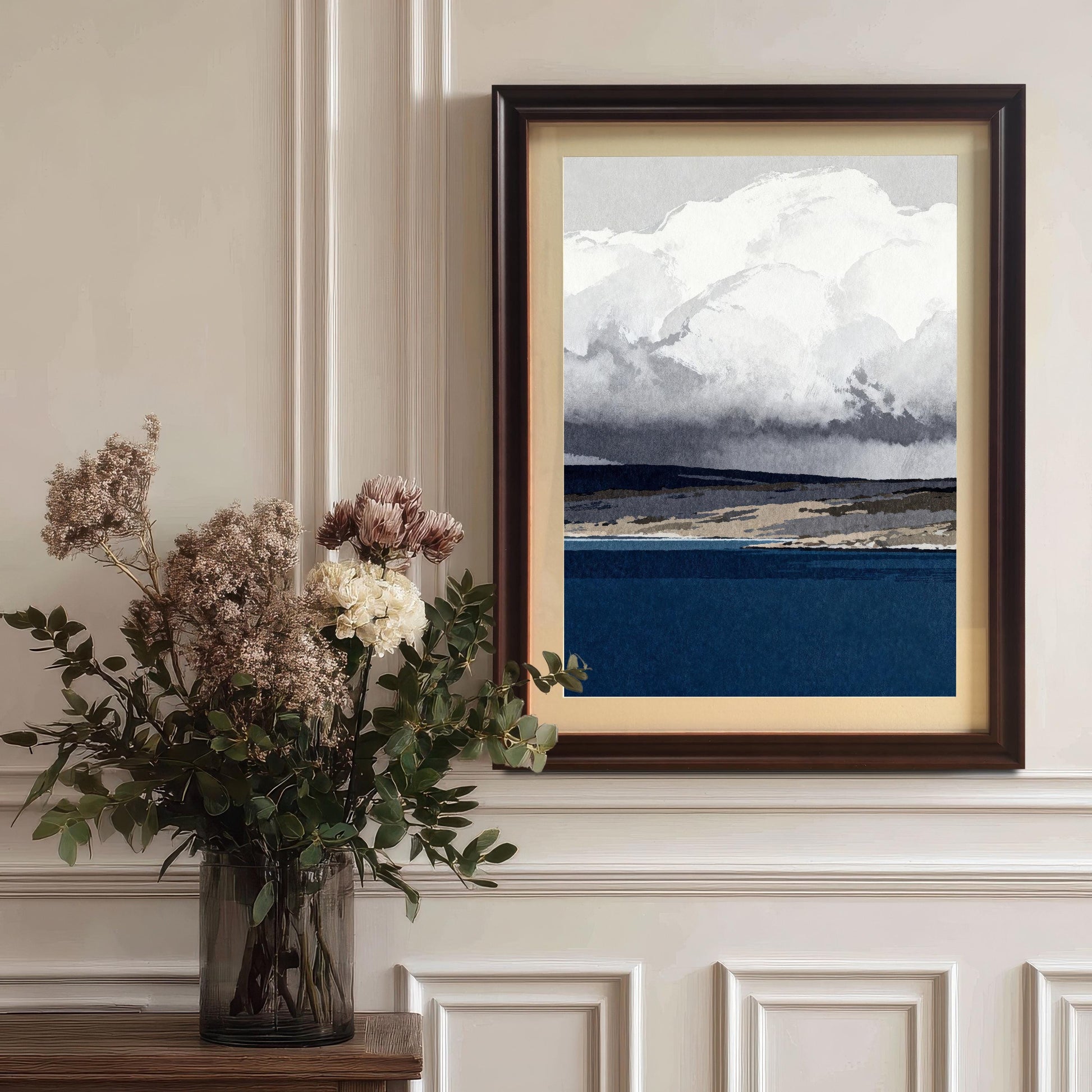 The abstract landscape art in a dark Coffee-colored wood frame with a cream mount, styled on a classic white wainscoted wall next to a vase of dried flowers.