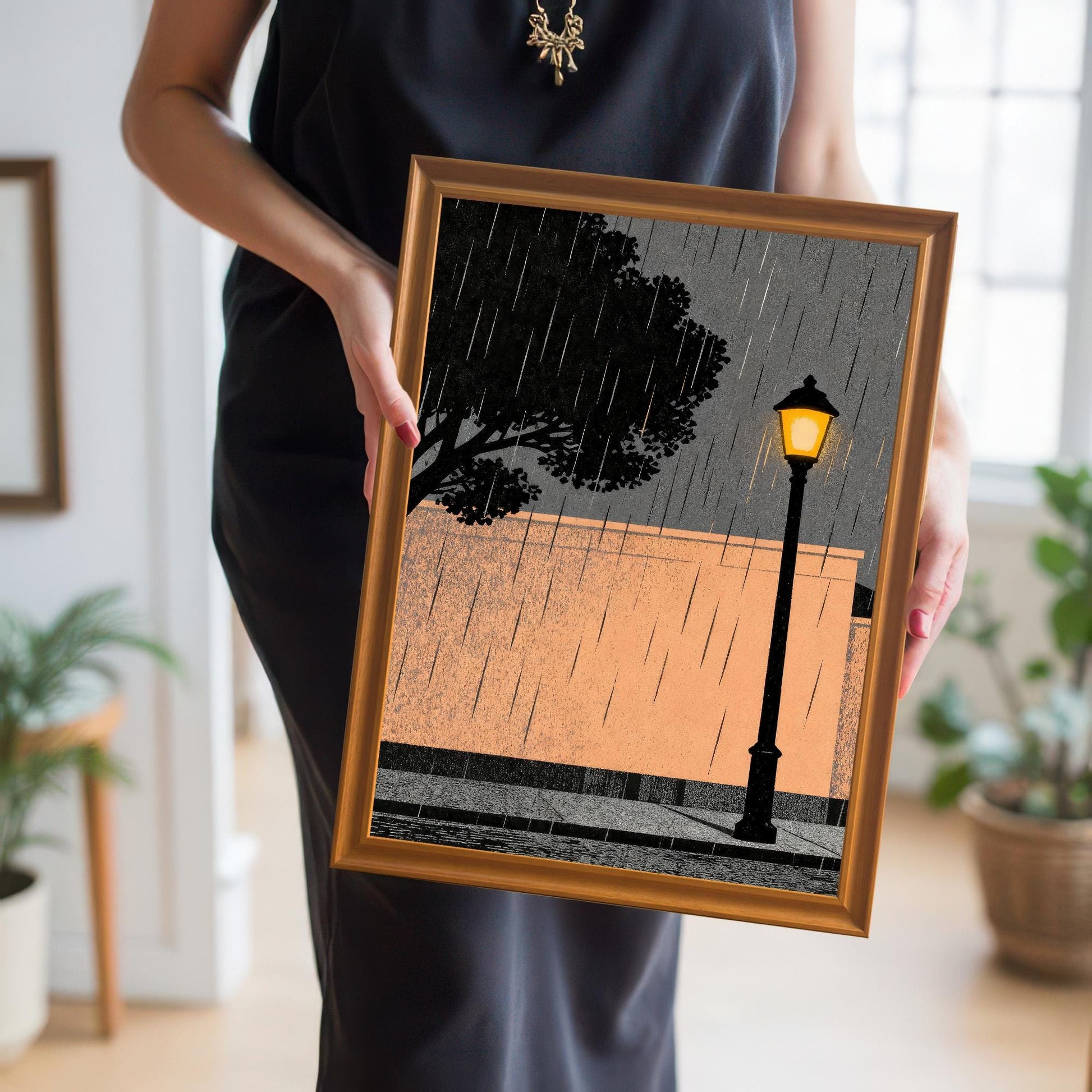 A person holding the medium-sized orange wood-framed art print, providing a real-world scale reference for the rainy street lamp illustration.