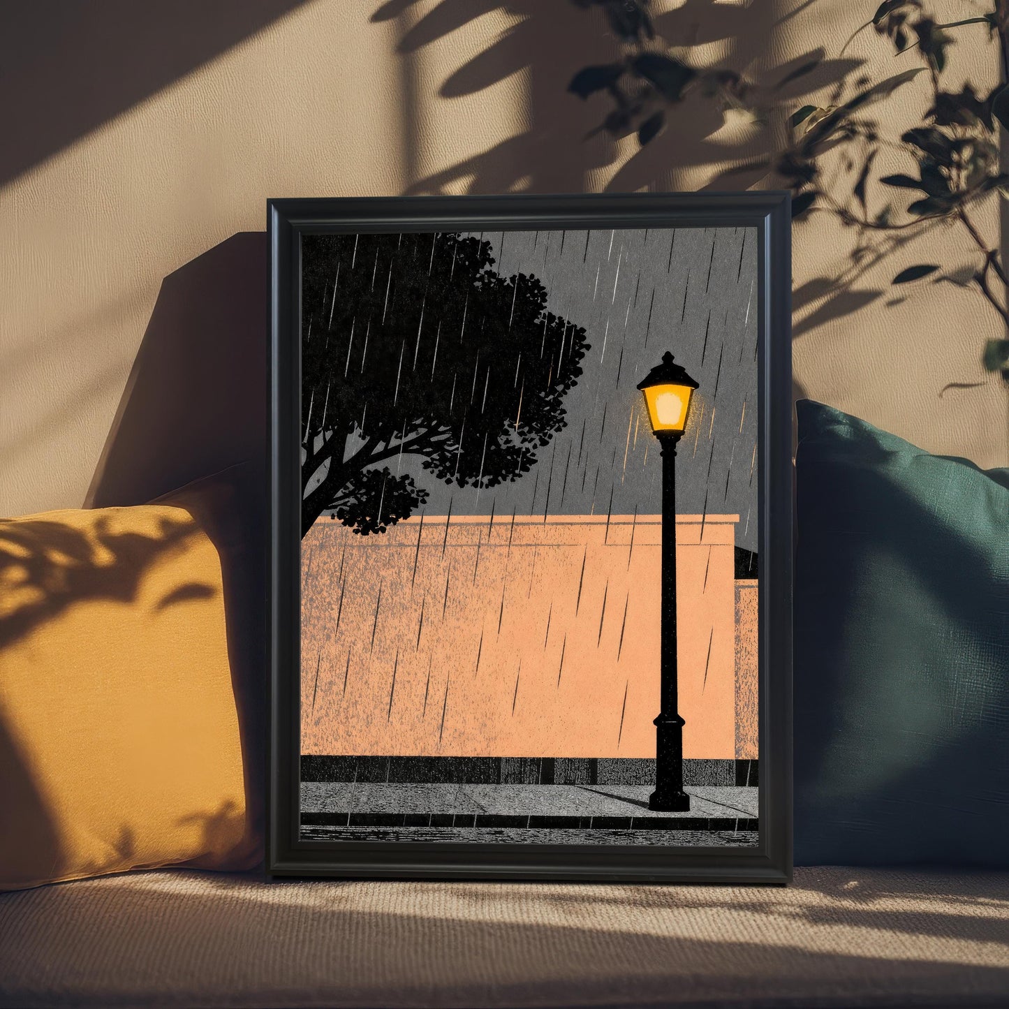 A black wood-framed art print of a rainy street scene resting on a textured surface between a yellow and dark green pillow, with soft leaf shadows cast across the wall and frame.