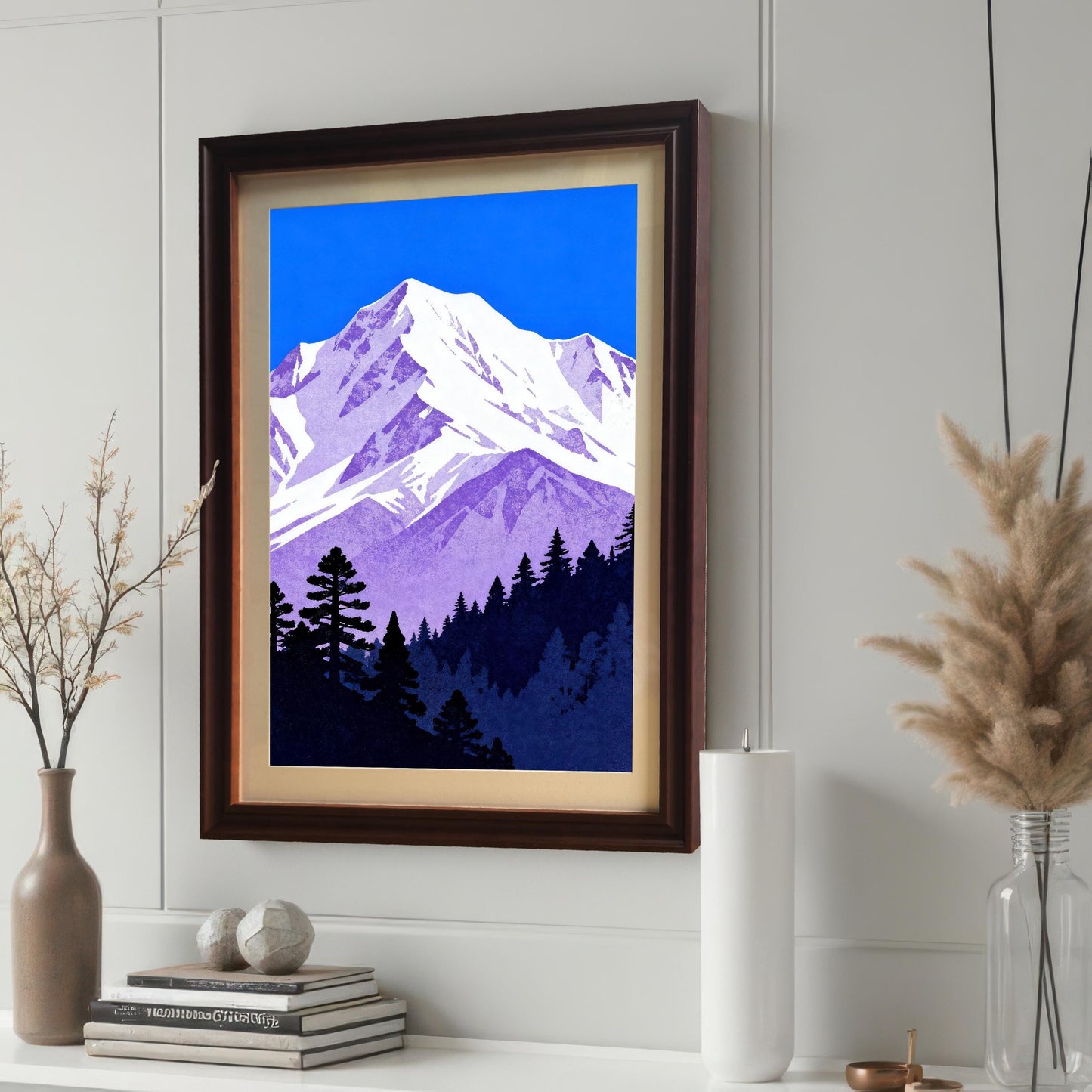 A purple and blue mountain landscape art print, framed in a dark brown 'Coffee' wood frame with a cream mount, displayed on a white wall above a stack of books and decor.