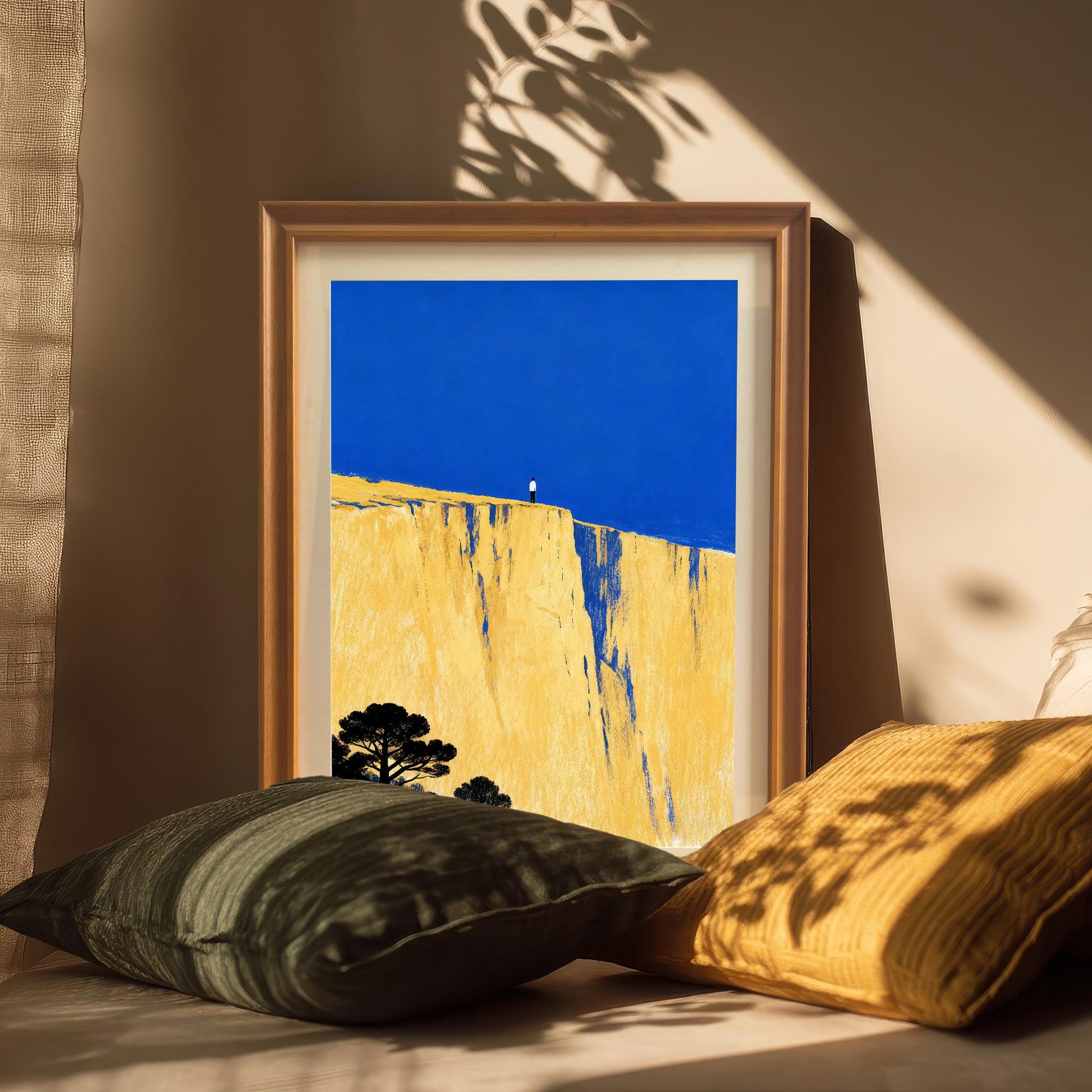 The art print in a light brown wood frame, resting on a beige surface surrounded by dark green and yellow cushions. Sunlight casts dramatic leaf and window shadows across the frame and background.