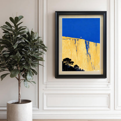 The framed art print featuring a figure on a yellow cliff beneath a blue sky, hanging on a white paneled wall next to a large potted Fiddle-leaf fig plant. The frame is black.