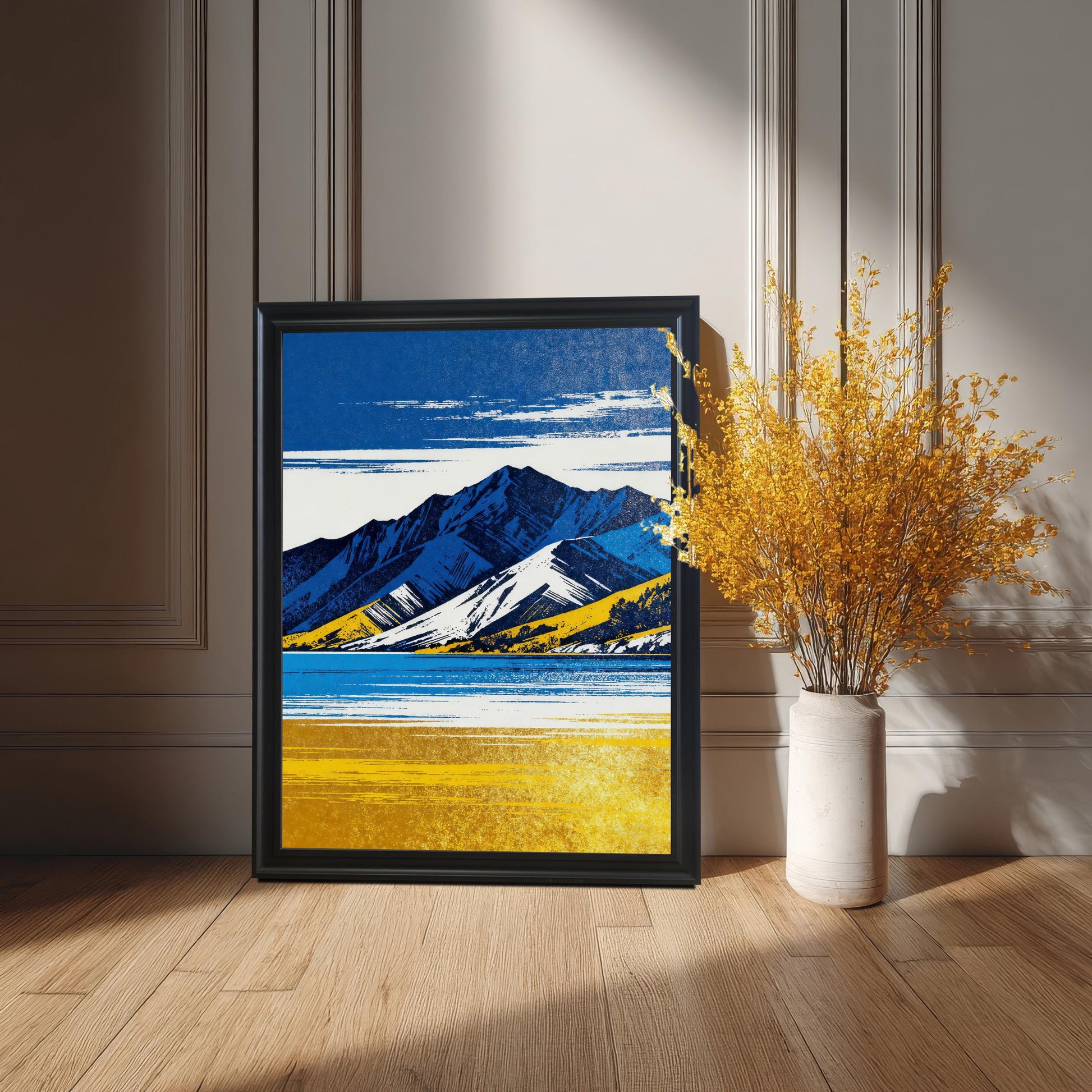 A framed art print in a black frame, featuring the blue and yellow mountain landscape, standing on a wooden floor against a gray paneled wall next to a tall white vase with yellow dried flowers.