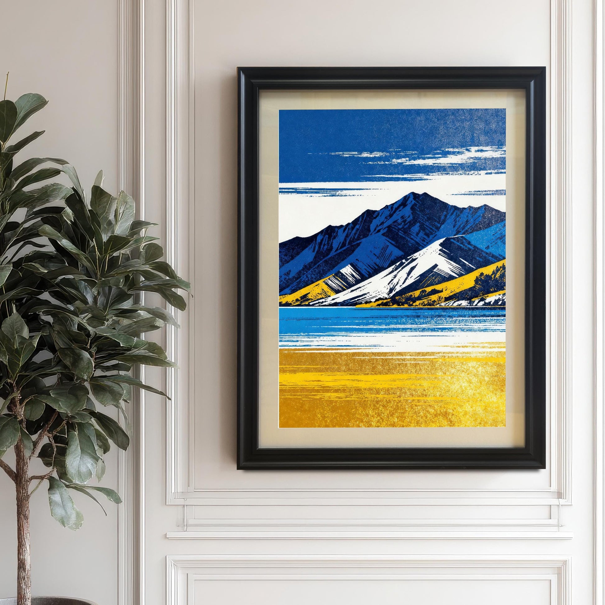 A framed art print in a black frame, featuring the blue and yellow mountain landscape, hanging on a white wall with wainscoting and trim. A large potted fiddle-leaf fig plant is situated to the left.
