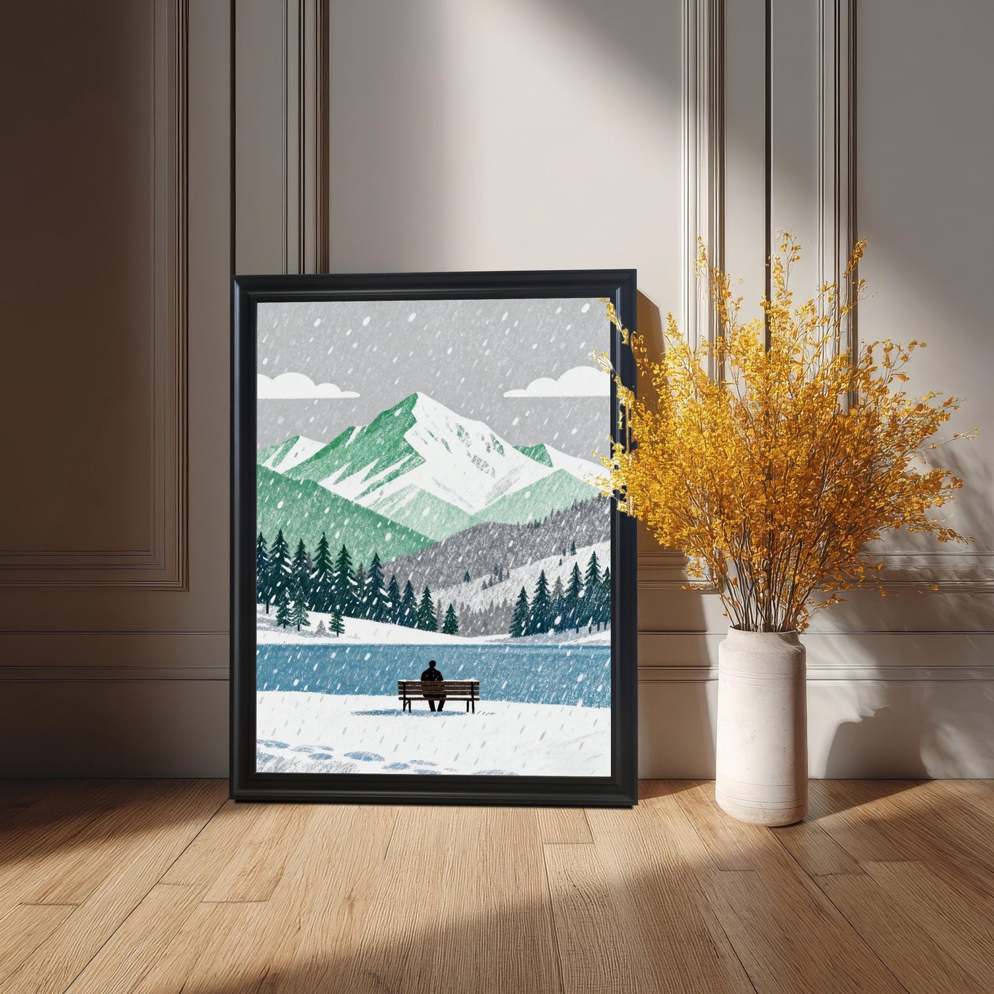 A framed art print in a black wood frame leaning against a paneled wall on a wooden floor, next to a white vase with tall, yellow dried flowers. The artwork features snowy mountains and a person on a bench.