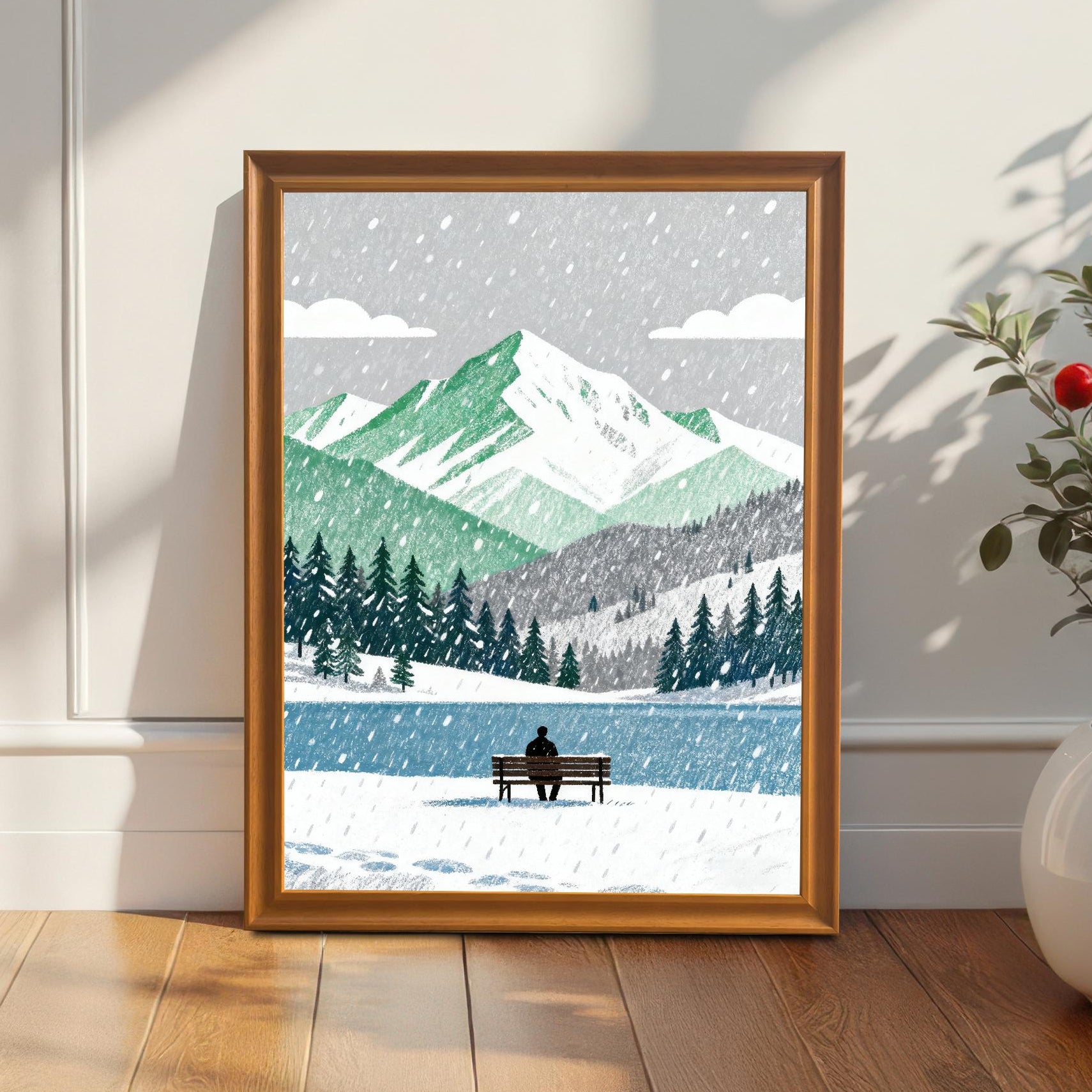 A framed art print in an orange wood frame leaning against a white paneled wall on a wooden floor, next to a white planter with a green plant and red berries. The artwork is a snowy mountain landscape.