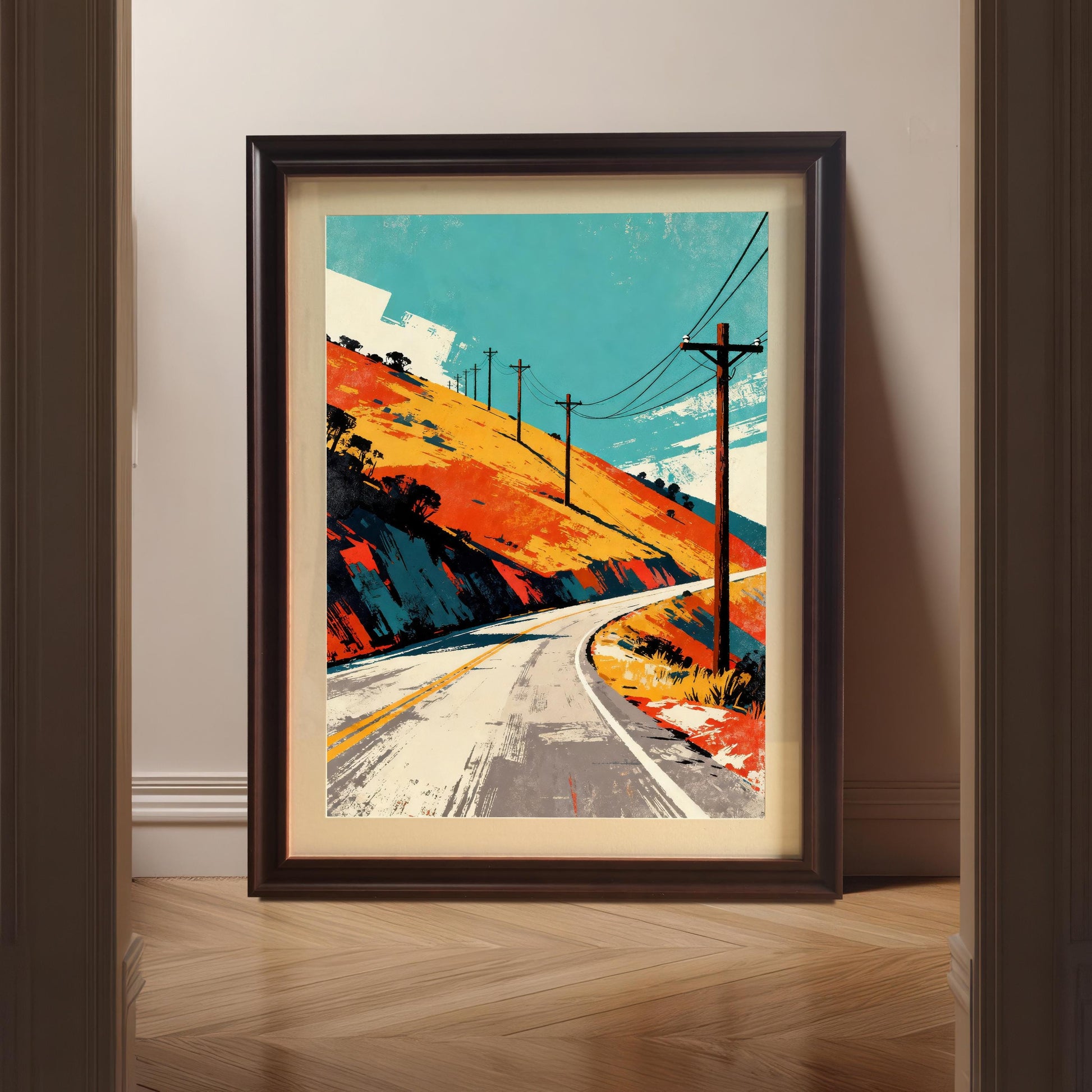 Framed art print leaning against a white wall on a wooden floor, viewed through a doorway. The print is a vibrant, abstract landscape of a curving road, orange hillside, and utility poles, in a dark wood or "Coffee" frame.