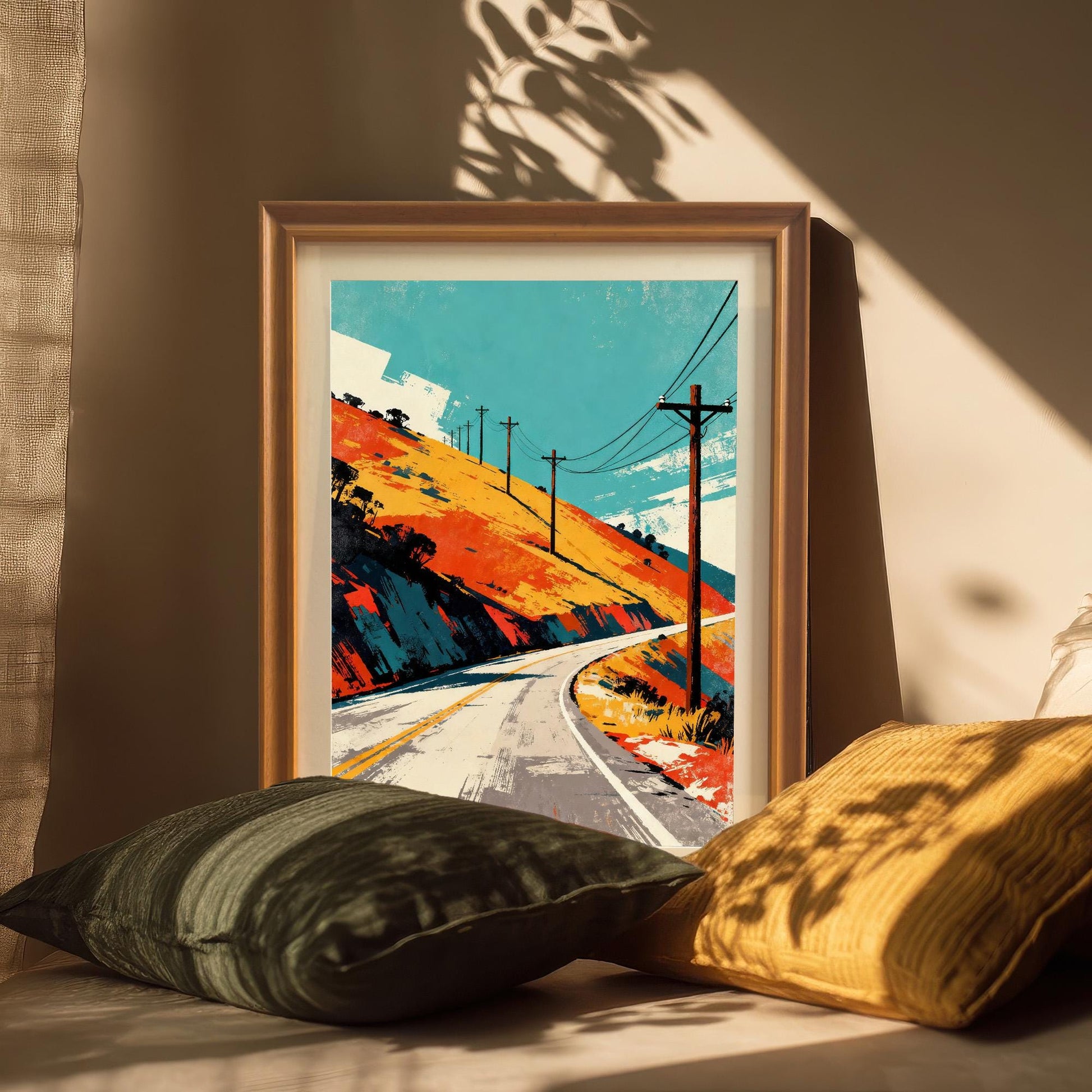 Framed art print in an Orange or light wood frame, leaning against a wall indoors, with two pillows in the foreground. Strong diagonal sunlight casts shadows of a nearby object (possibly leaves or a plant) over the print and background.