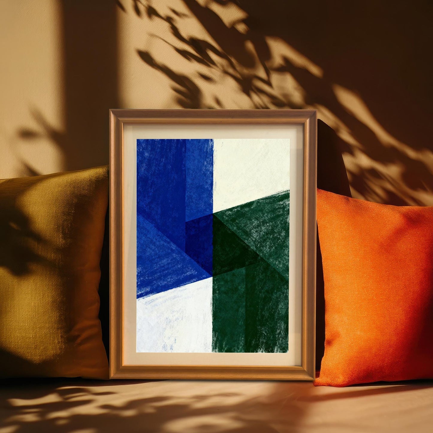 Orange-framed abstract art print with a white mat, resting between two colorful pillows (mustard yellow and bright orange). The artwork features bold geometric shapes in blue, green, and cream white. Sunlight casts leaf shadows across the scene.