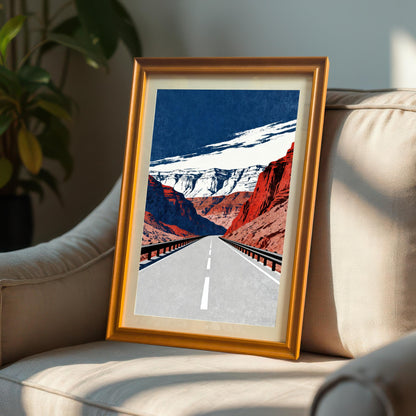 A framed art print with a bright gold/orange frame, leaning on the arm of a light-colored armchair in a sunny indoor setting. The art depicts a long road through canyons.