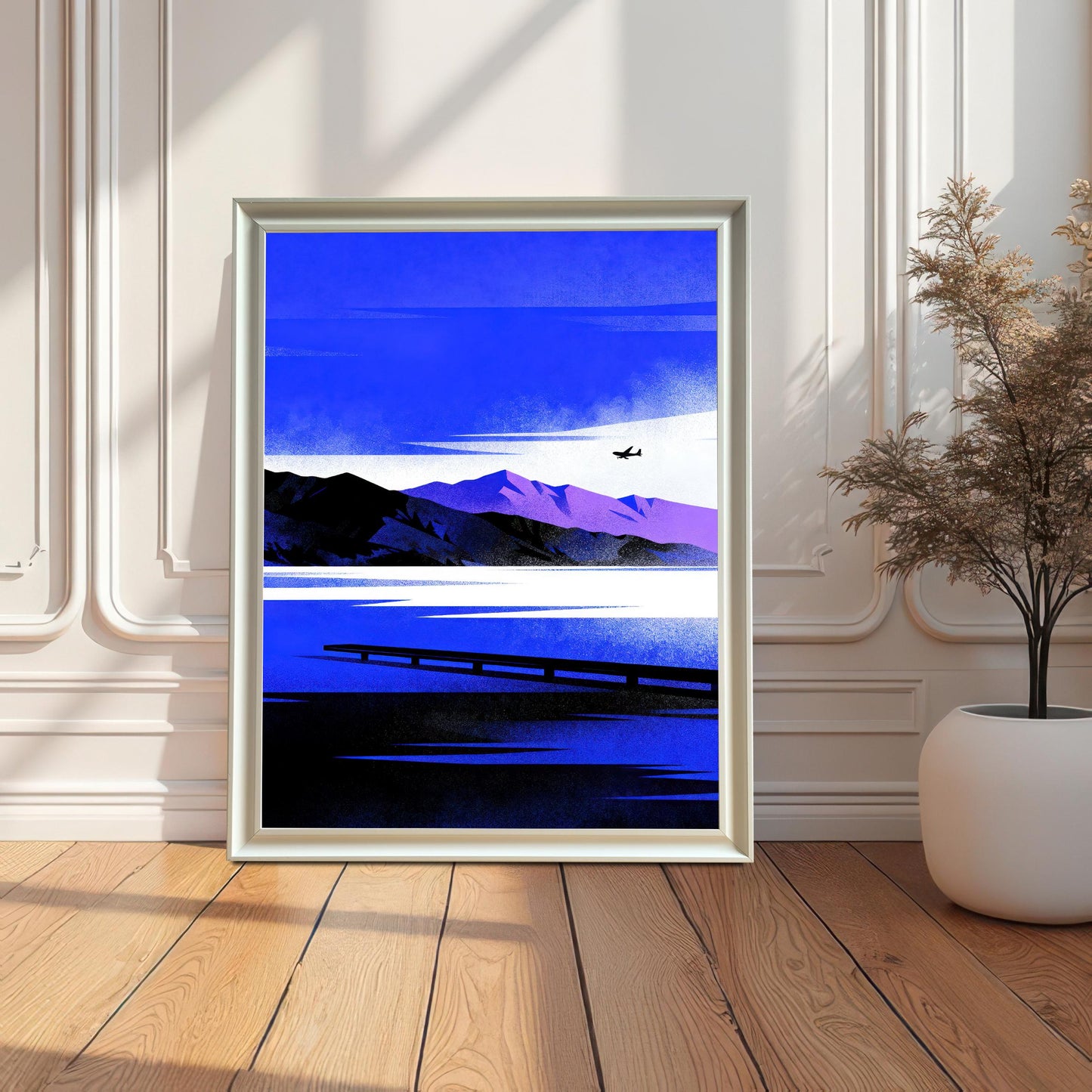 White-framed art print leaning on a light wood floor against a white wall with decorative molding. The artwork shows a stylized landscape in deep blue, white, black, and purple, depicting a lake, distant mountains, a pier, and a small airplane. A dried plant in a white pot is on the right.