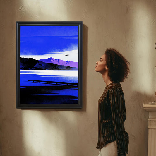 A woman with curly hair, wearing a striped shirt, standing in profile and looking up at the framed art print hanging on a textured beige wall. The artwork is a modern landscape illustration in bold blues and purple, depicting a lake, mountains, and a small airplane.