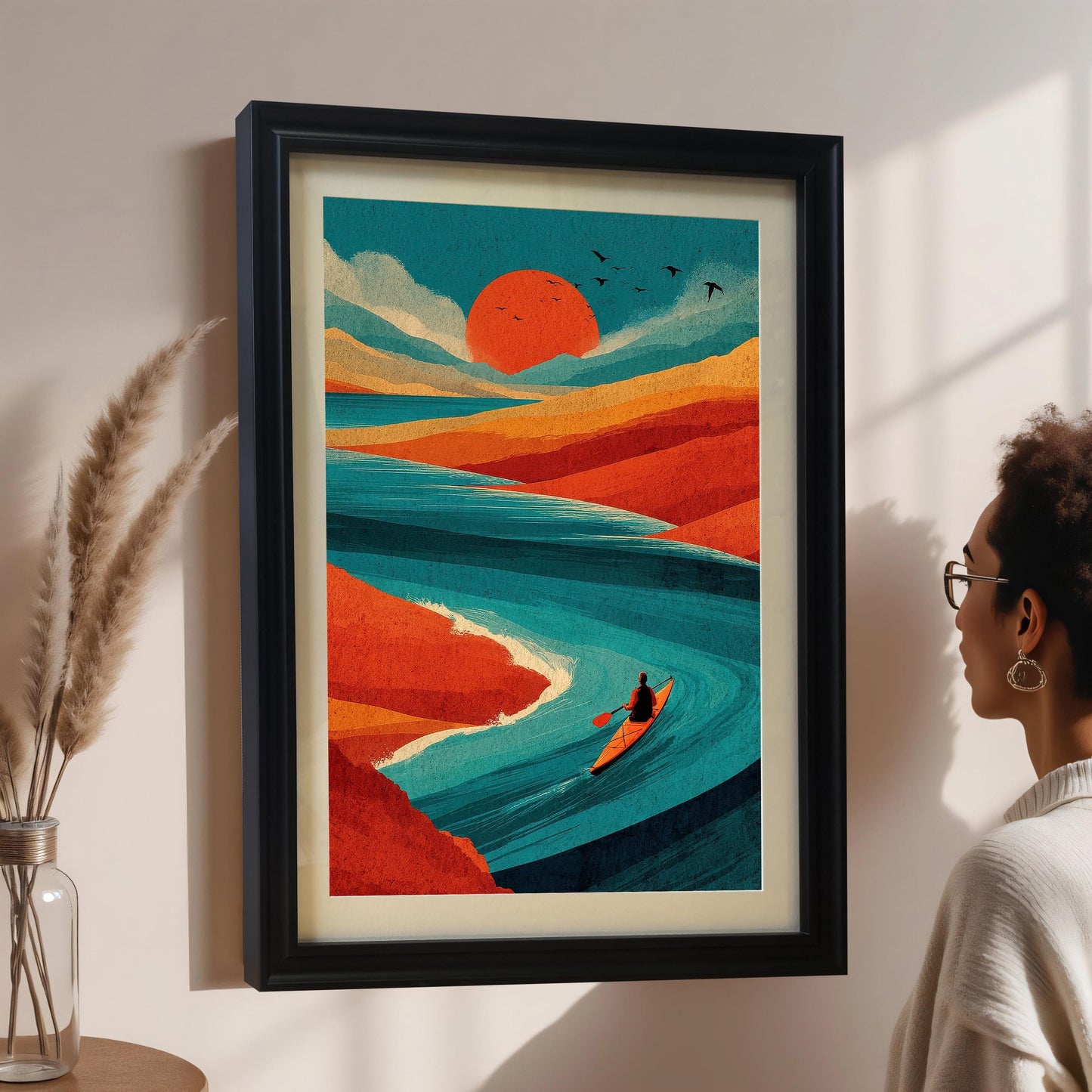 A person viewing a large black-framed landscape art print of a teal river and red sun, mounted on a neutral wall in a brightly lit room.