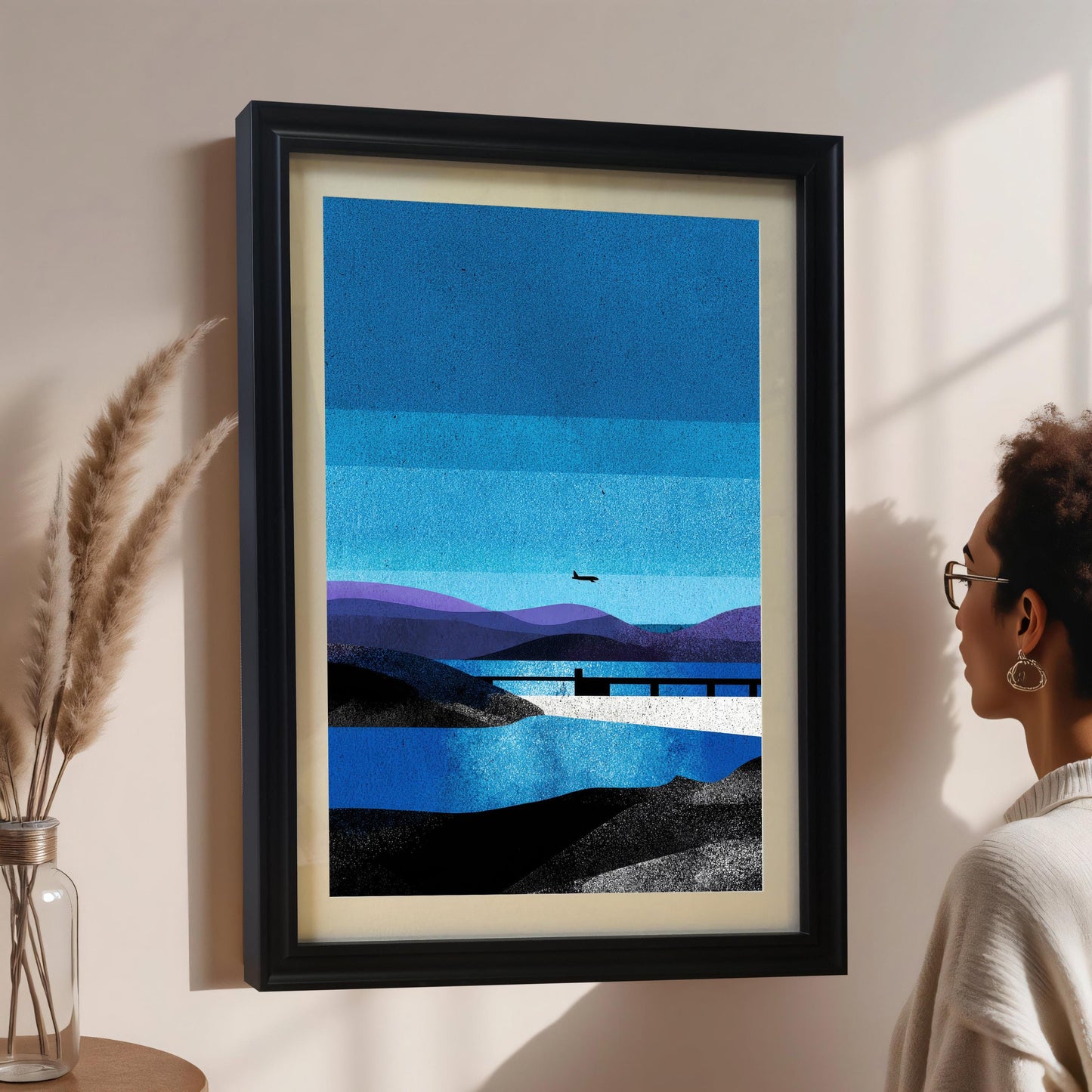 A lifestyle shot showing a black wood-framed art print with a cream mount hanging on a neutral wall; a person in profile and a vase of dried pampas grass are in the foreground.