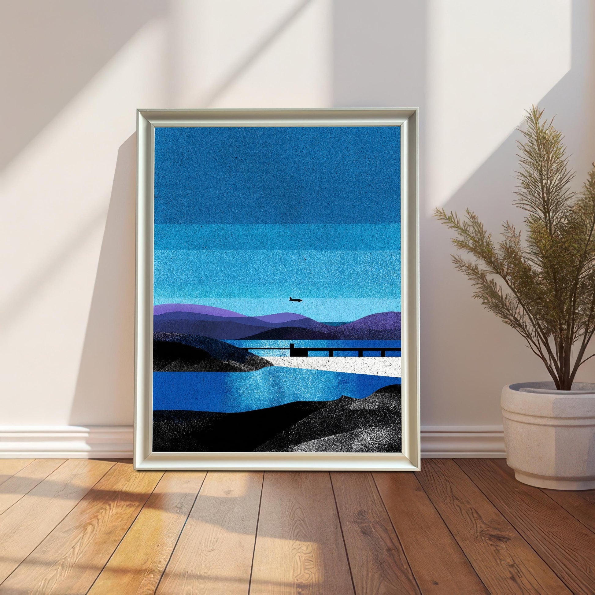 A full-bleed "White" wood-framed art print sitting on a wooden floor against a white wall, positioned next to a potted green plant in a bright, sunlit room.