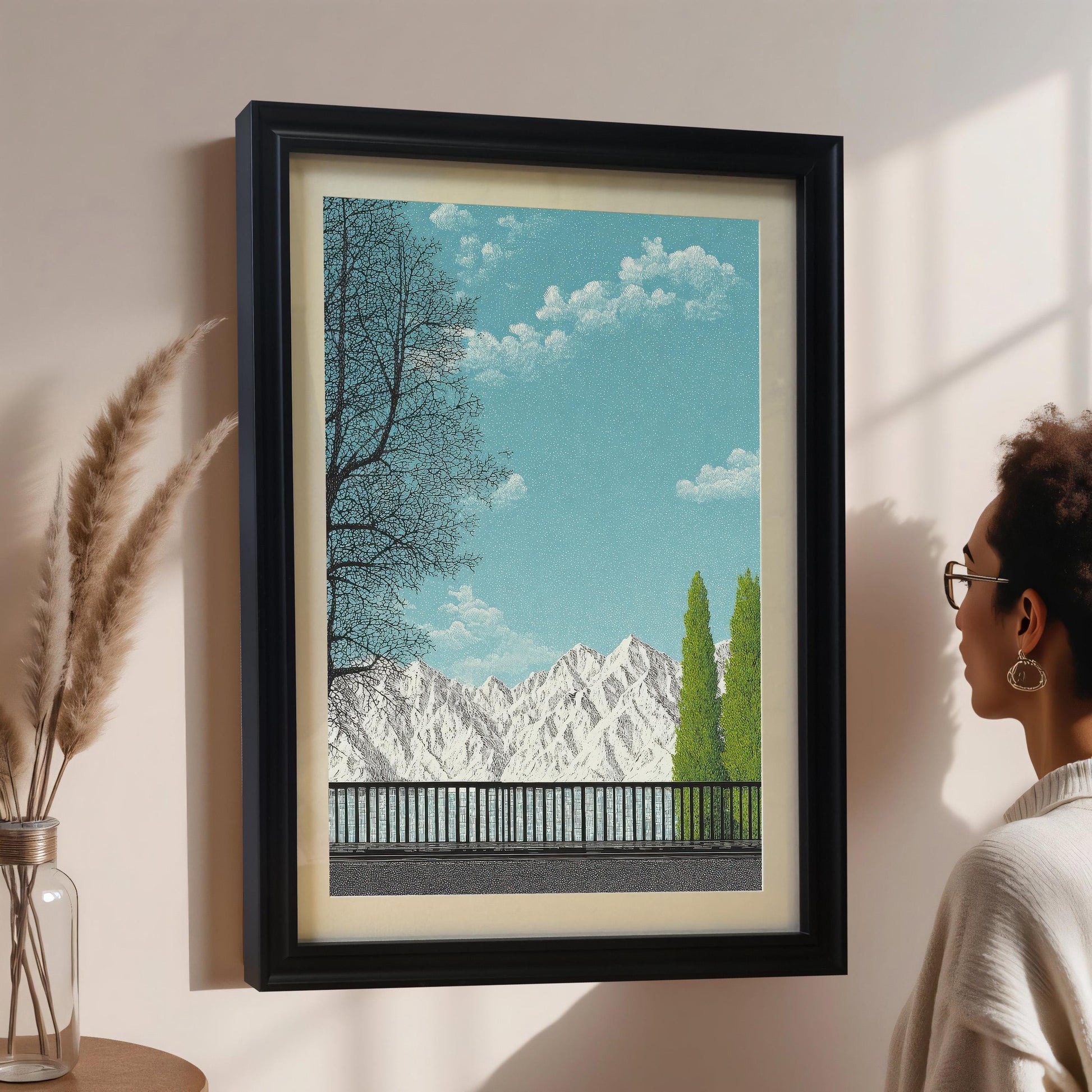 A side-view mockup of a Black wood framed art print with a white mount, displayed on a neutral wall next to a person and a decorative vase with dried pampas grass.