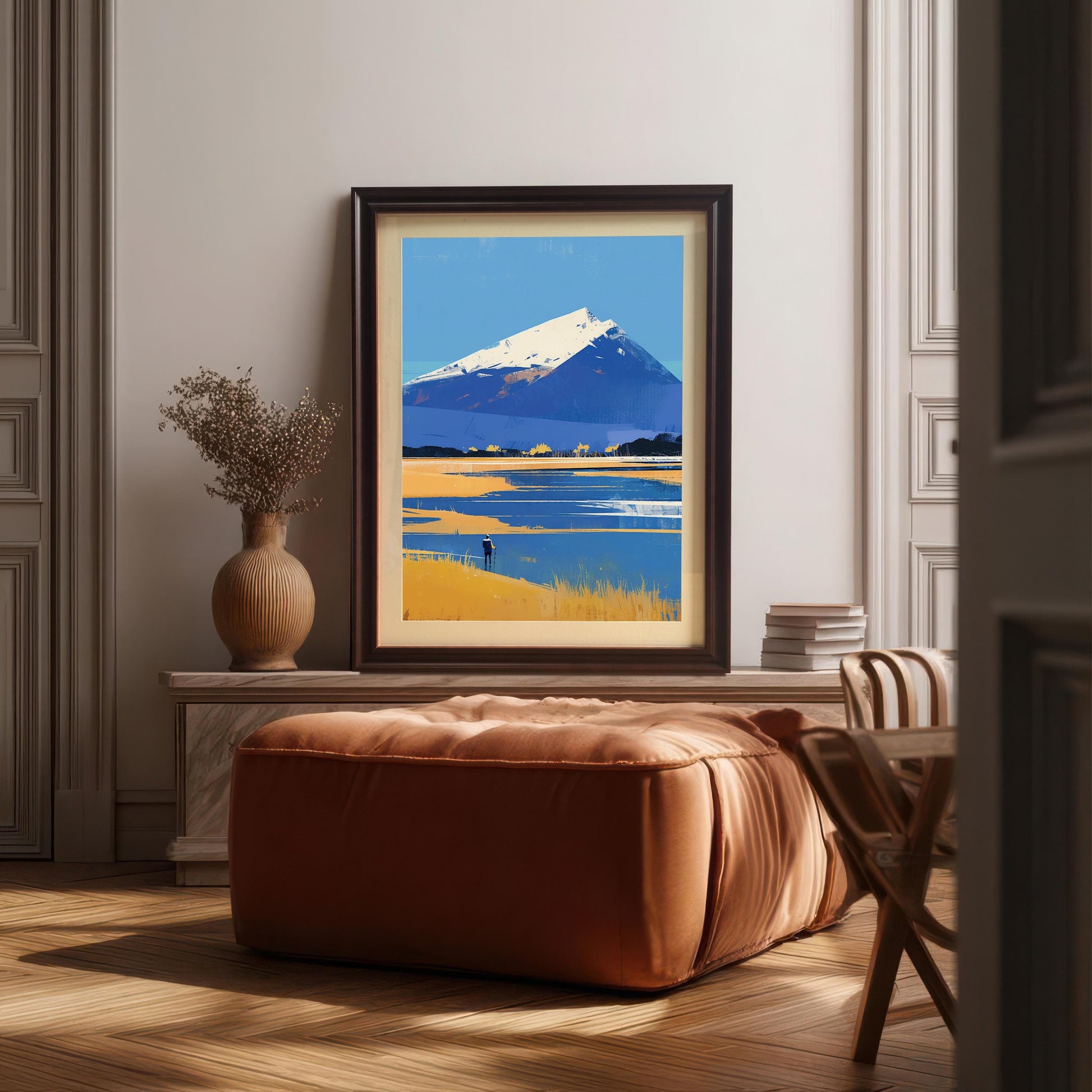The mountain landscape artwork in a dark wood frame with a mount, leaning on a marble-topped sideboard in an elegant room. In front of the art is a large, square cognac-colored leather ottoman, and to the left is a textured ceramic vase holding dried decorative branches.