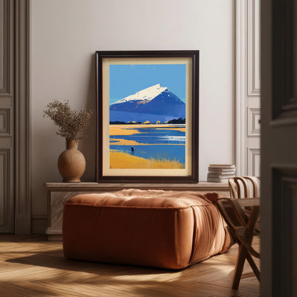 The mountain landscape artwork in a dark wood frame with a mount, leaning on a marble-topped sideboard in an elegant room. In front of the art is a large, square cognac-colored leather ottoman, and to the left is a textured ceramic vase holding dried decorative branches.