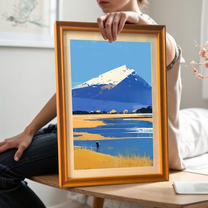 A person holding the mountain art print in an orange wood frame with a mount. The person is sitting on a wooden surface, wearing dark jeans, providing a real-world scale reference for the framed piece.