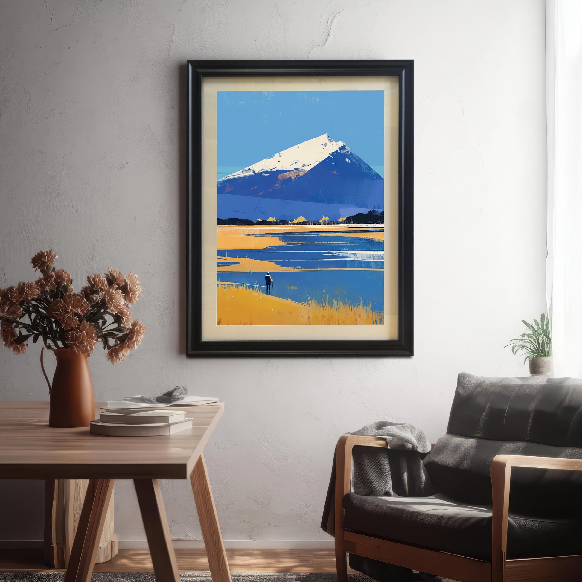 A large mountain landscape art print in a sleek black frame with an off-white mount, hanging on a light gray textured wall. The scene is set in a modern living room featuring a dark leather chair with a gray throw blanket and a wooden table with a vase of dried flowers in the foreground.