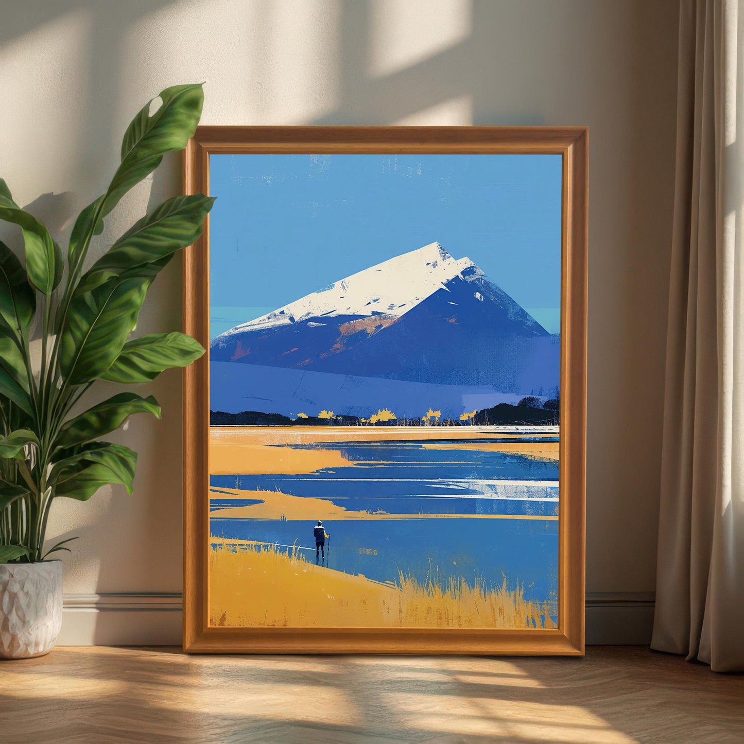 The mountain landscape art in a honey-toned wood frame, leaning against a neutral-colored wall on a wooden floor. A large green leafy plant is to the left, and a beige curtain is to the right, with warm sunlight filtering into the room.