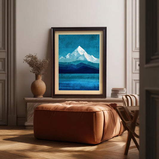A dark brown wood-framed mountain art print with a cream-colored mount, resting on a stone mantelpiece in a sophisticated room. The scene features a plush orange leather ottoman in the foreground, a vase of dried flowers, and sunlight streaming across a classic parquet floor.
