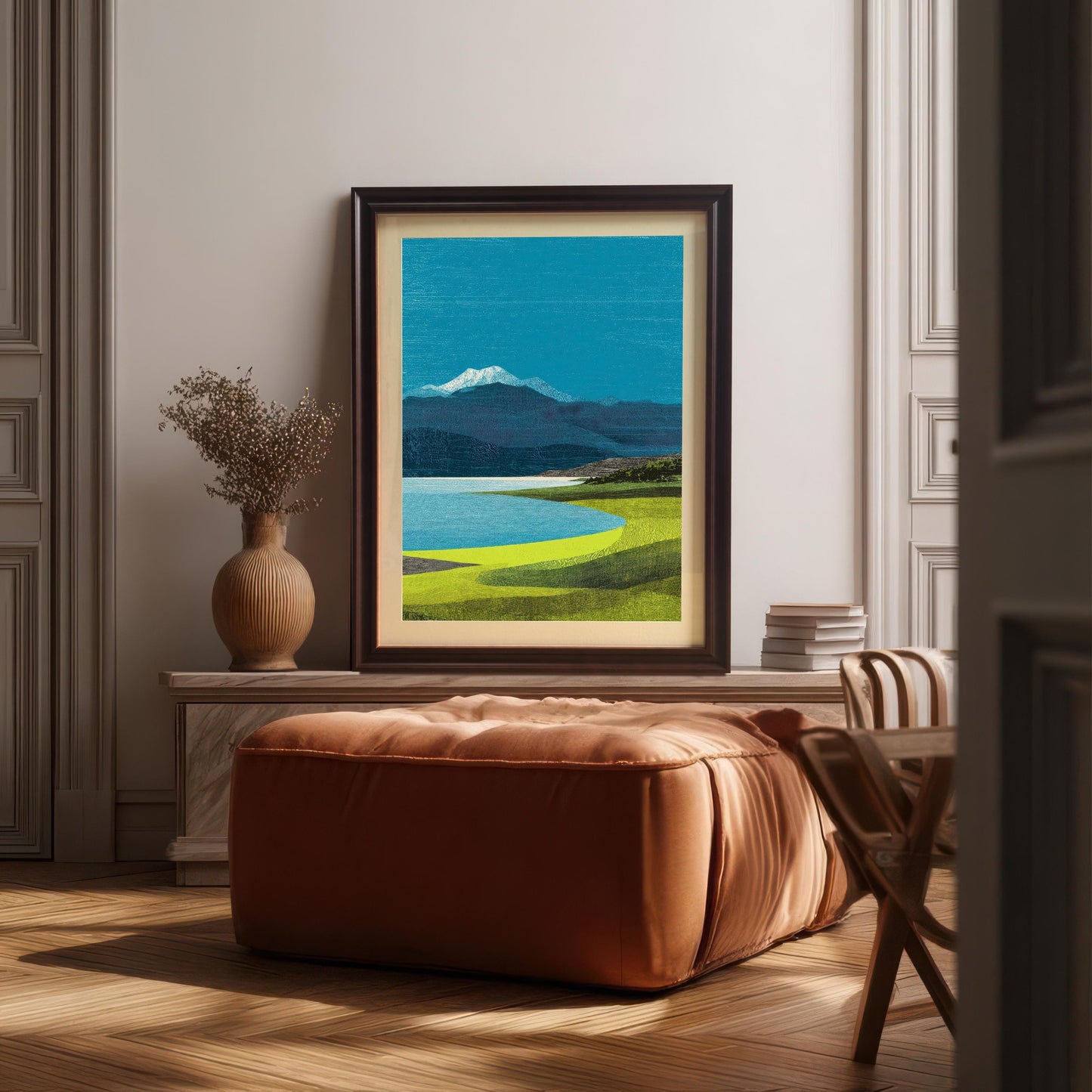 A landscape art print in a coffee-colored wood frame with a mount, styled on a marble console table behind a large leather ottoman in a sunlit room.