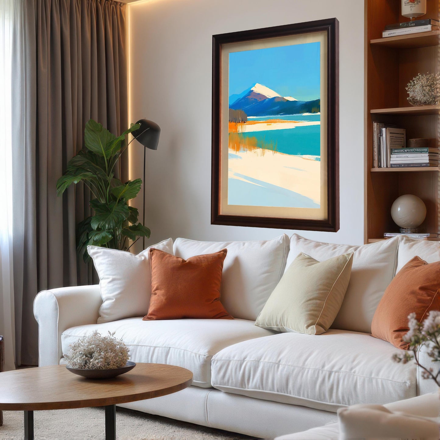 A mountain landscape art print with a dark coffee-colored frame and cream mount hanging above a white sectional sofa. The living room is decorated with orange and beige throw pillows, a round wooden coffee table, and a built-in bookshelf.
