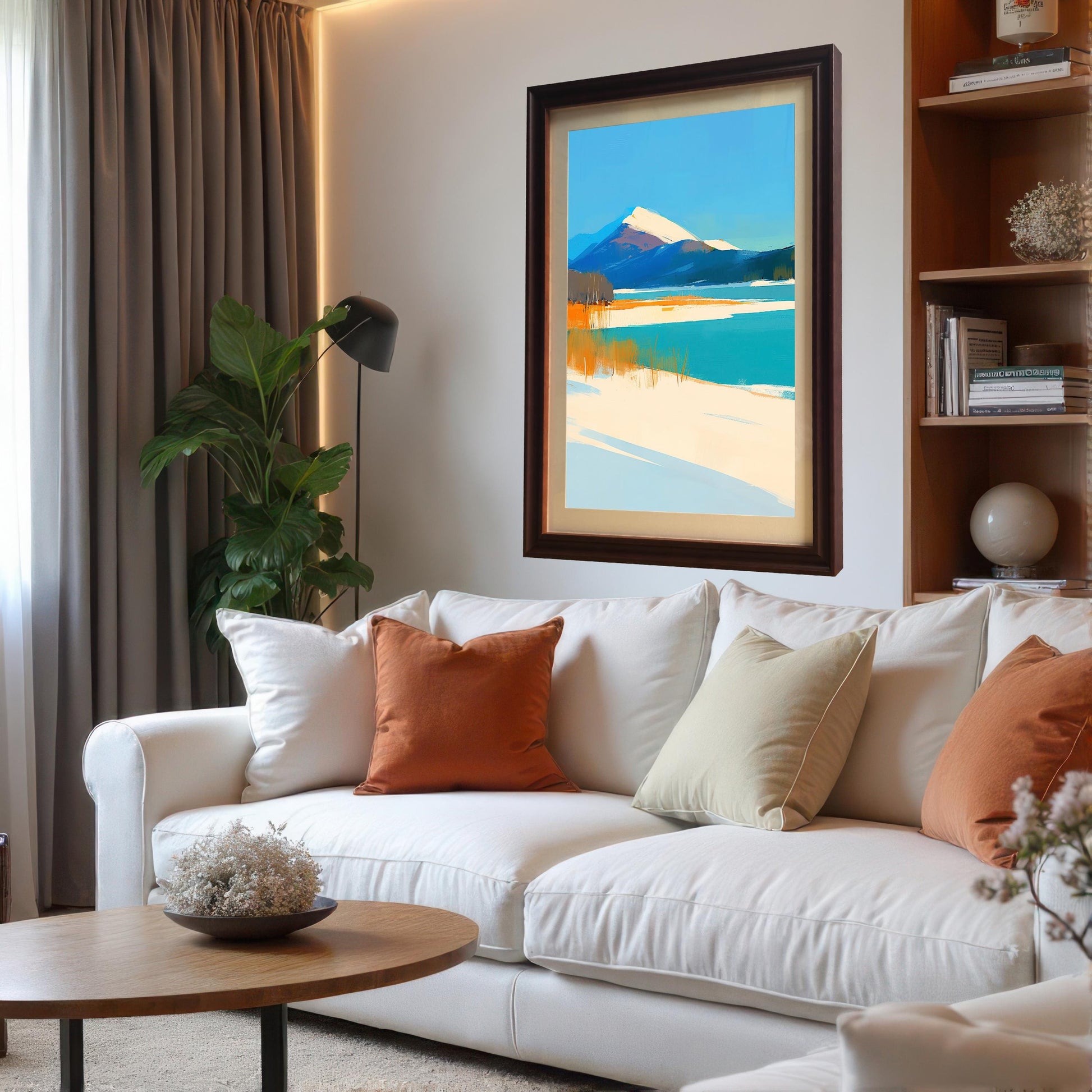 A mountain landscape art print with a dark coffee-colored frame and cream mount hanging above a white sectional sofa. The living room is decorated with orange and beige throw pillows, a round wooden coffee table, and a built-in bookshelf.